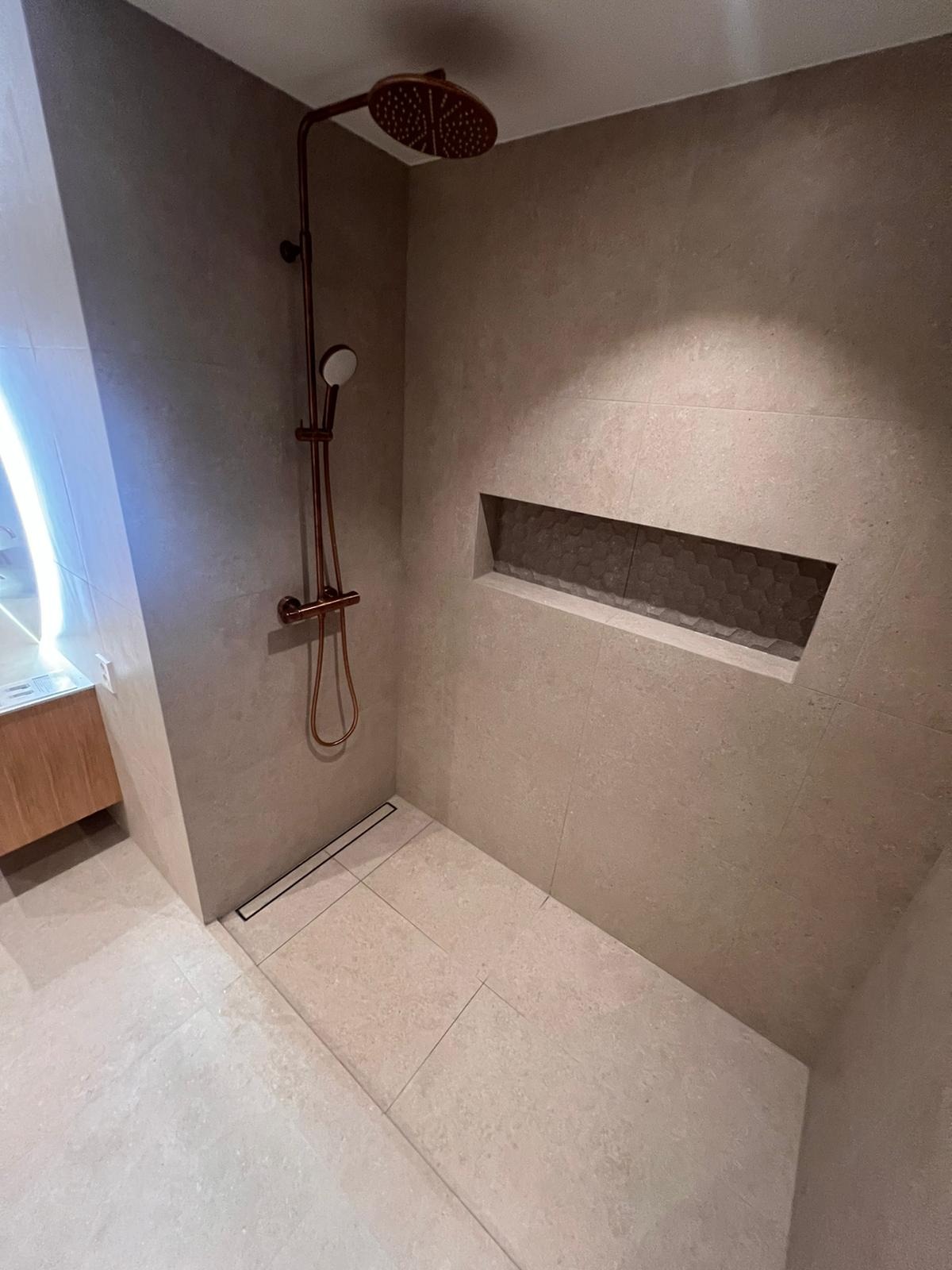 Nowoczesna kabina prysznicowa z deszczownicą w kolorze miedzi, beżowe płytki wielkoformatowe, odpływ liniowy i wnęka w ścianie wykończona heksagonalnymi płytkami.