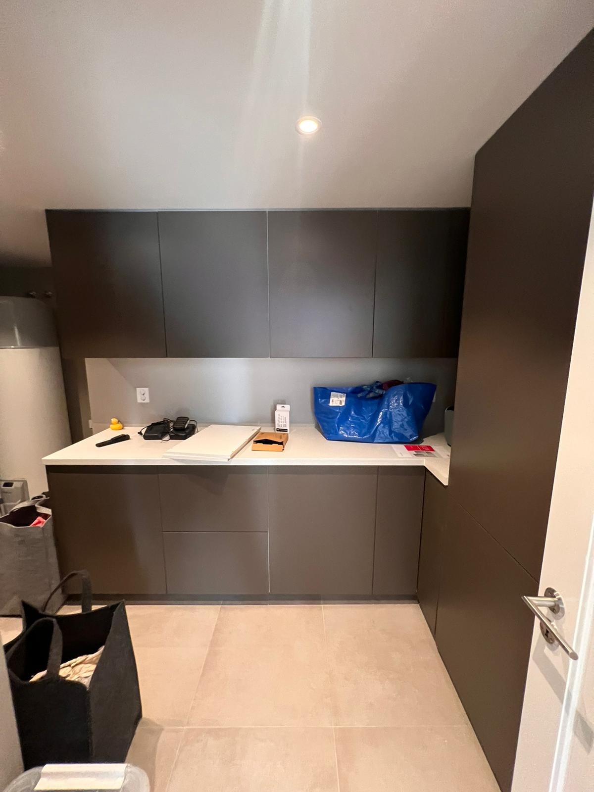 Nowoczesna łazienka z szarymi szafkami, białym blatem, beżowymi płytkami podłogowymi i niebieską torbą na blacie; widoczne drobne przedmioty i akcesoria.