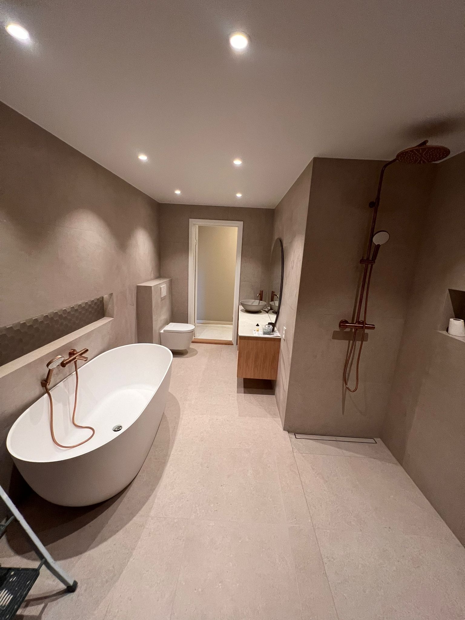 Nowoczesna łazienka z wolnostojącą wanną, armaturą w kolorze miedzi, beżowymi ścianami, umywalką na drewnianej szafce, toaletą w zabudowie i prysznicem typu walk-in z odpływem liniowym.