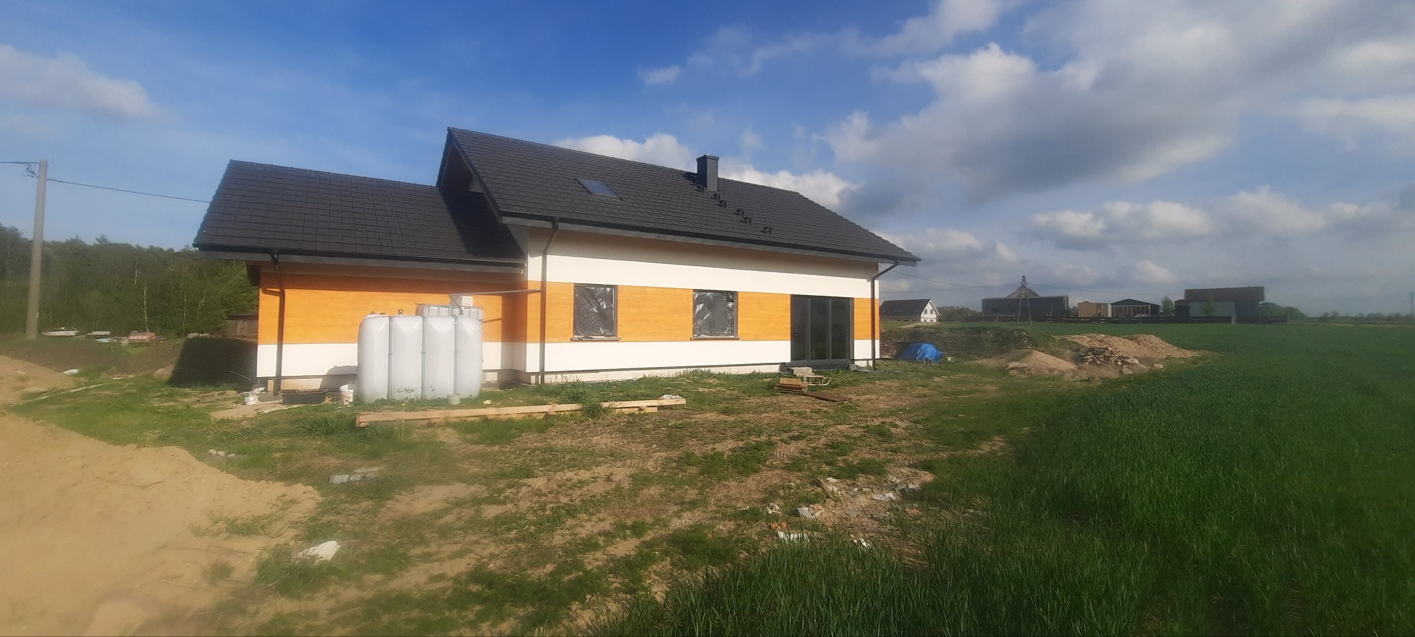 Nowo wybudowany dom jednorodzinny z czarnym dachem, elewacją w kolorze białym i pomarańczowym, okna zabezpieczone folią, widoczne zbiorniki na wodę przy ścianie, trawnik z przodu, budynki...