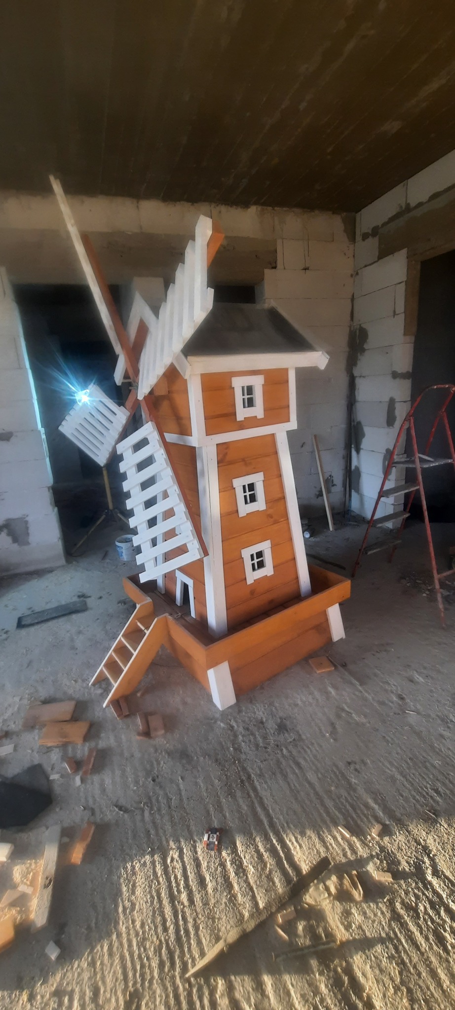 Drewniany model wiatraka w pomarańczowo-białych barwach, z mini schodkami, umieszczony w surowym wnętrzu w trakcie remontu, z widoczną drabiną i porozrzucanymi cegłami na podłodze.