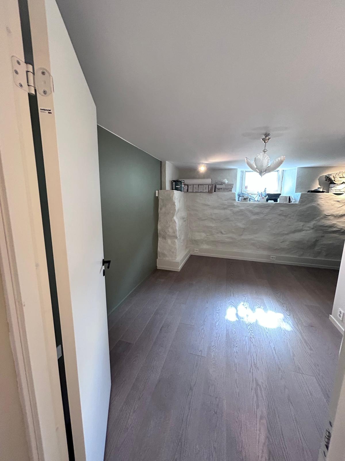 Wnętrze pokoju z szarobrązową podłogą z paneli, fragmentem ściany pomalowanej na oliwkowy kolor oraz ścianą wyłożoną kamieniem w kolorze białym, widok zza otwartych drzwi.