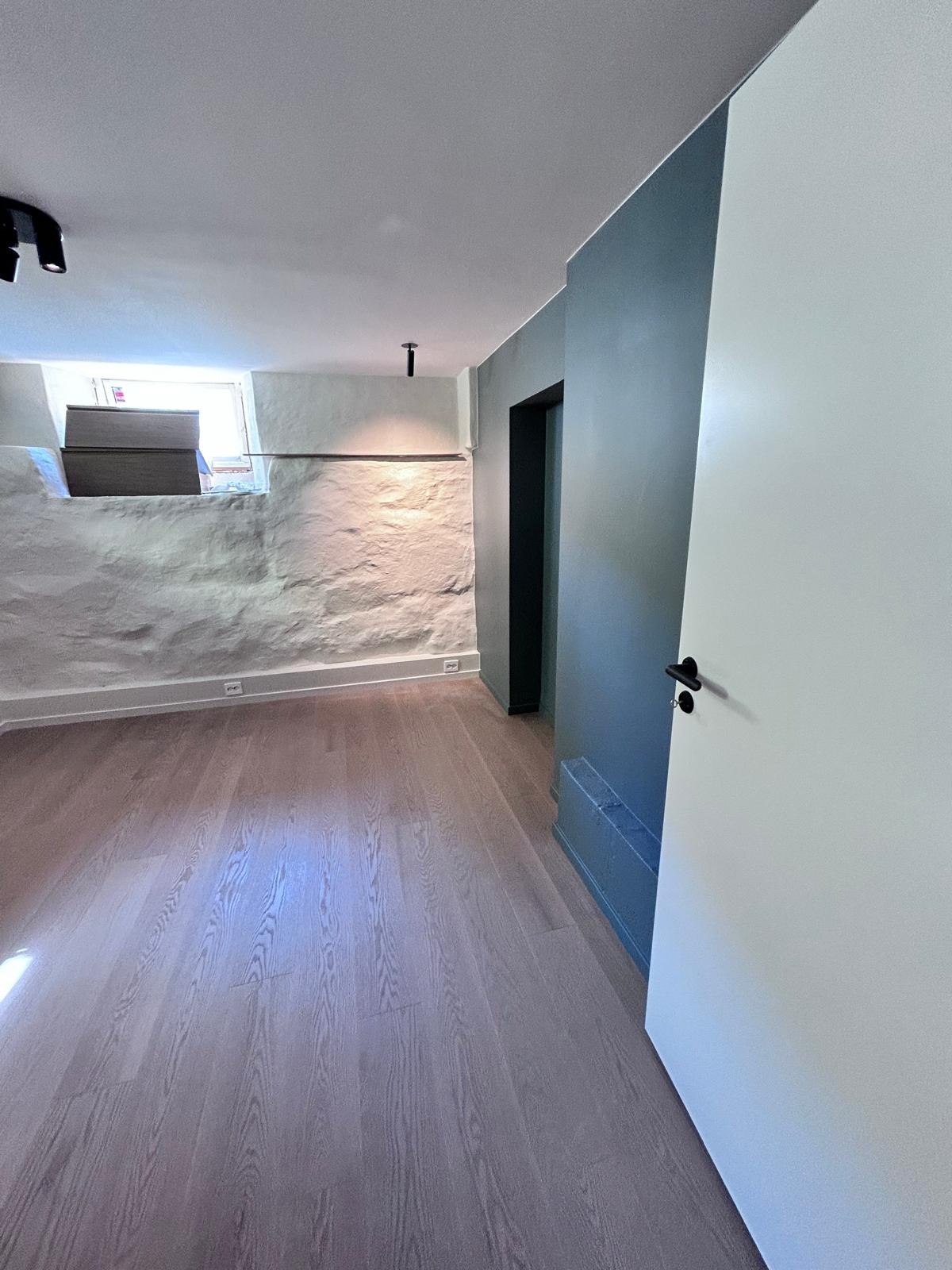 Wnętrze po odnowieniu: jasna podłoga z paneli, szara ściana z surowego kamienia, błękitna ściana z ukrytym grzejnikiem i białe drzwi uchylone do środka.