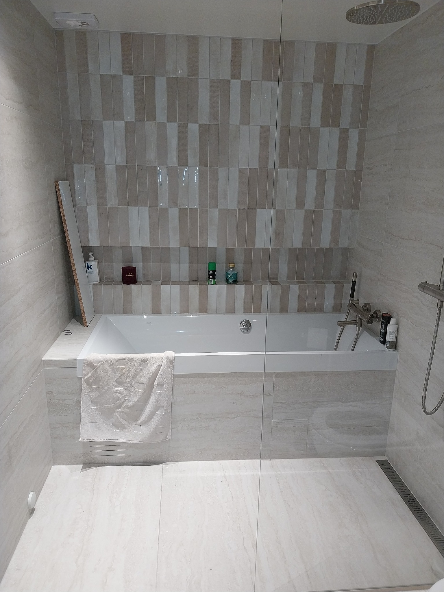 Nowoczesna łazienka z wanną obłożoną beżowymi płytkami imitującymi marmur, szklany parawan nawannowy, deszczownica i półka w ścianie z kosmetykami.