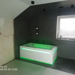 Nowoczesna wanna z hydromasażem, podświetlona na zielono, w łazience z szarą ścianą i oknem, widoczne ślady prac wykończeniowych na ścianie po lewej.