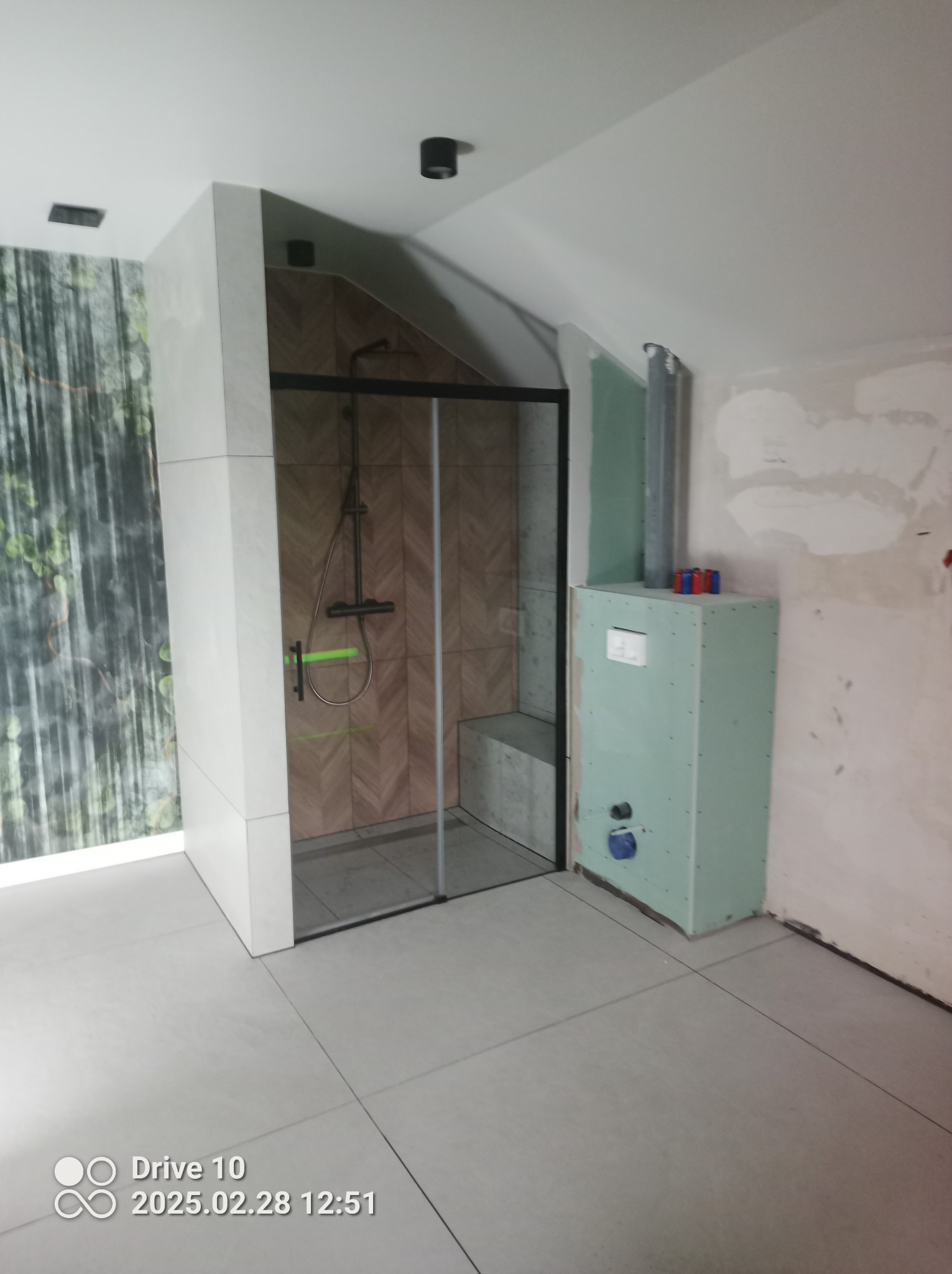 Wnętrze łazienki w trakcie remontu: kabina prysznicowa z czarnymi profilami i siedziskiem, wykończona płytkami w jodełkę, obok zabudowa z płyty gipsowej z instalacją sanitarną, fragment ściany...