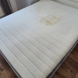 Biały materac na łóżku z widoczną plamą, na tle drewnianej podłogi i białej szafy.