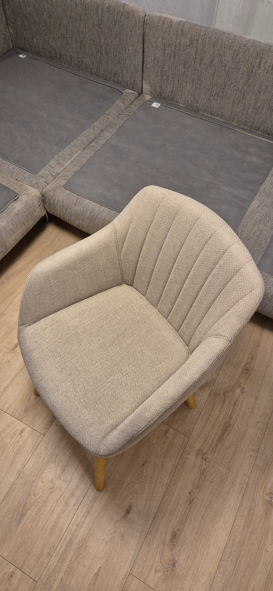Beżowy fotel z tapicerowanym siedziskiem i oparciem, ustawiony na drewnianej podłodze, z fragmentem narożnika sofy w tle.