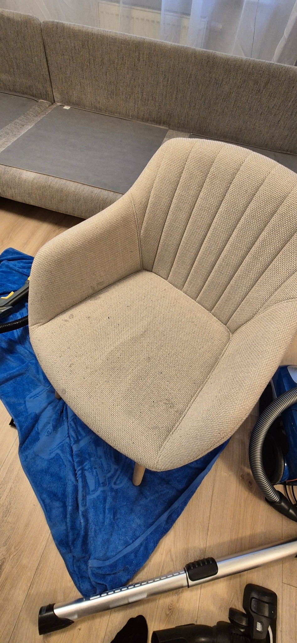 Beżowe krzesło tapicerowane z widocznymi plamami na siedzisku, leżące na niebieskim ręczniku obok odkurzacza, sugerujące przygotowanie do czyszczenia.
