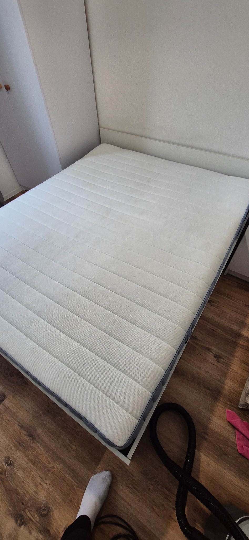 Świeżo wyprany, biały materac z regularnymi przeszyciami, leżący na białej ramie łóżka, z widocznym wężem odkurzacza piorącego i fragmentem skarpetki na drewnianej podłodze.