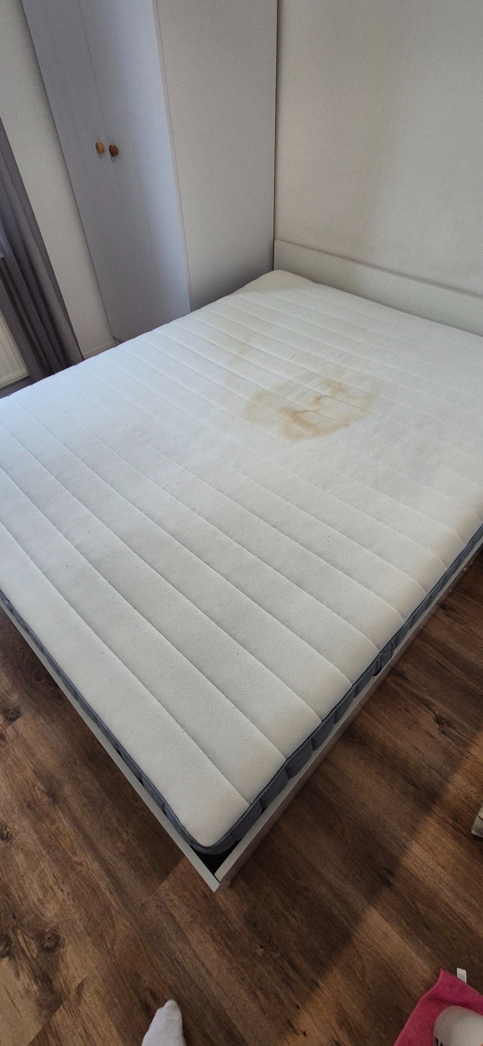 Biały materac na łóżku z widoczną plamą, na tle drewnianej podłogi i białej szafy.