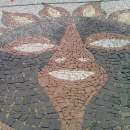 Mozaika z kostki brukowej przedstawiająca schematyczny wizerunek twarzy, wykonana z czarnej, białej, szarej i czerwonej kostki, widoczna z góry.