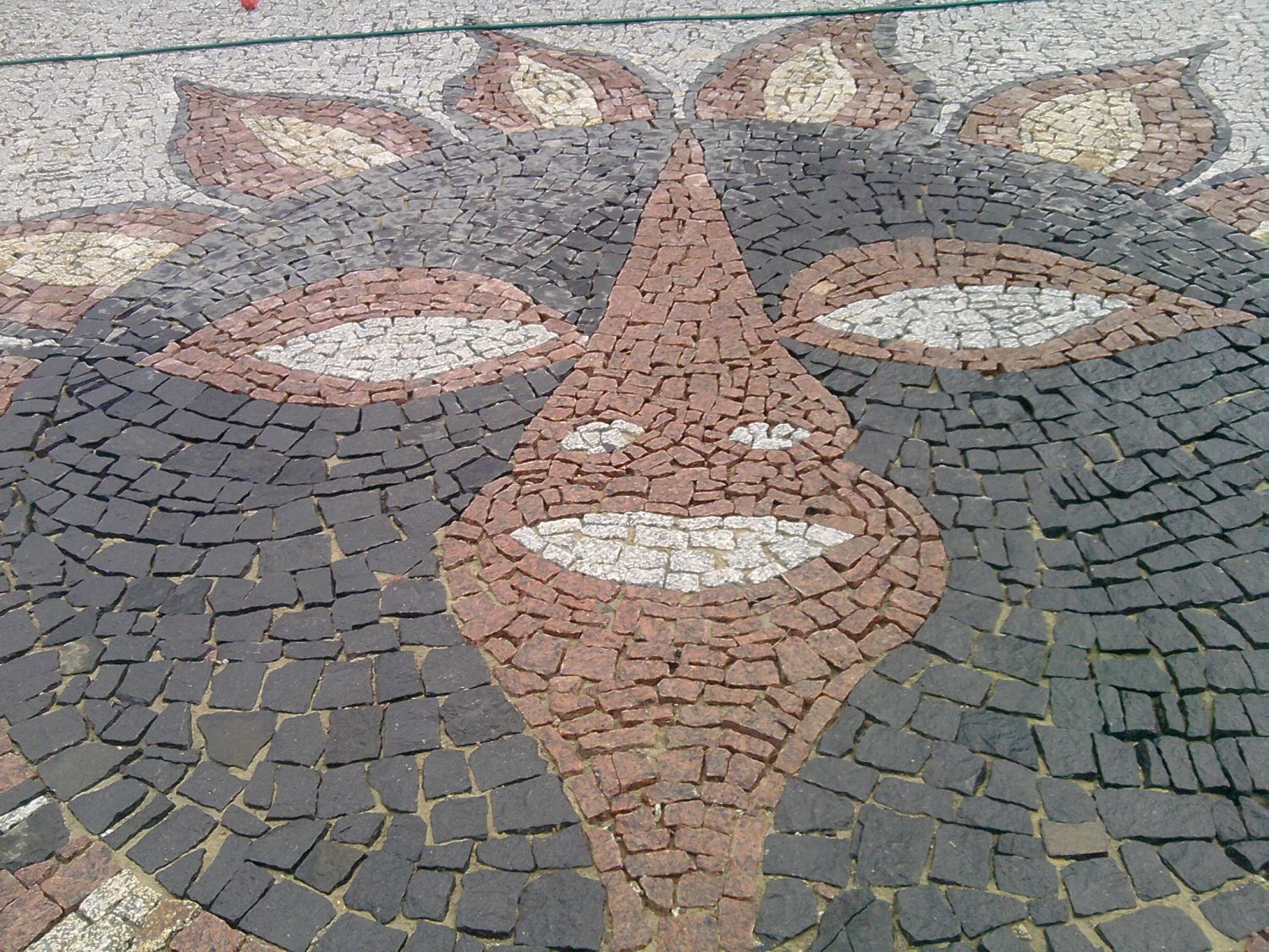 Mozaika z kostki brukowej przedstawiająca schematyczny wizerunek twarzy, wykonana z czarnej, białej, szarej i czerwonej kostki, widoczna z góry.