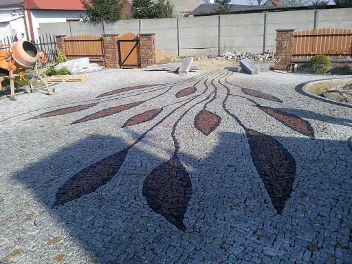 Ogród z ścieżką z jasnoszarej kostki, zdobioną wzorem liści z ciemniejszej kostki, widoczny betonowy mur i brama w tle, oraz betoniarka po lewej stronie.