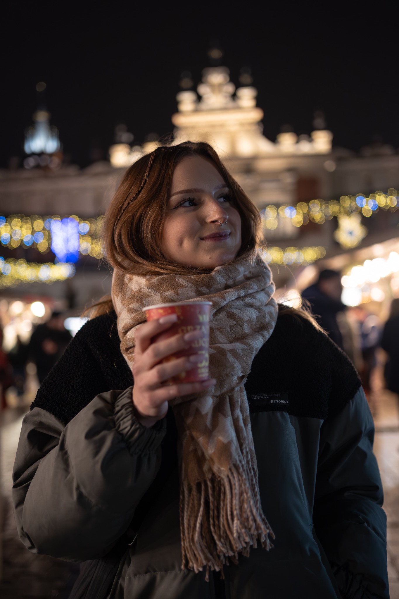 Uśmiechnięta kobieta z kolczykiem w nosie, trzymająca kubek z napojem na tle rozświetlonego Rynku Głównego w Krakowie nocą, ubrana w zimową kurtkę i wzorzysty szal.