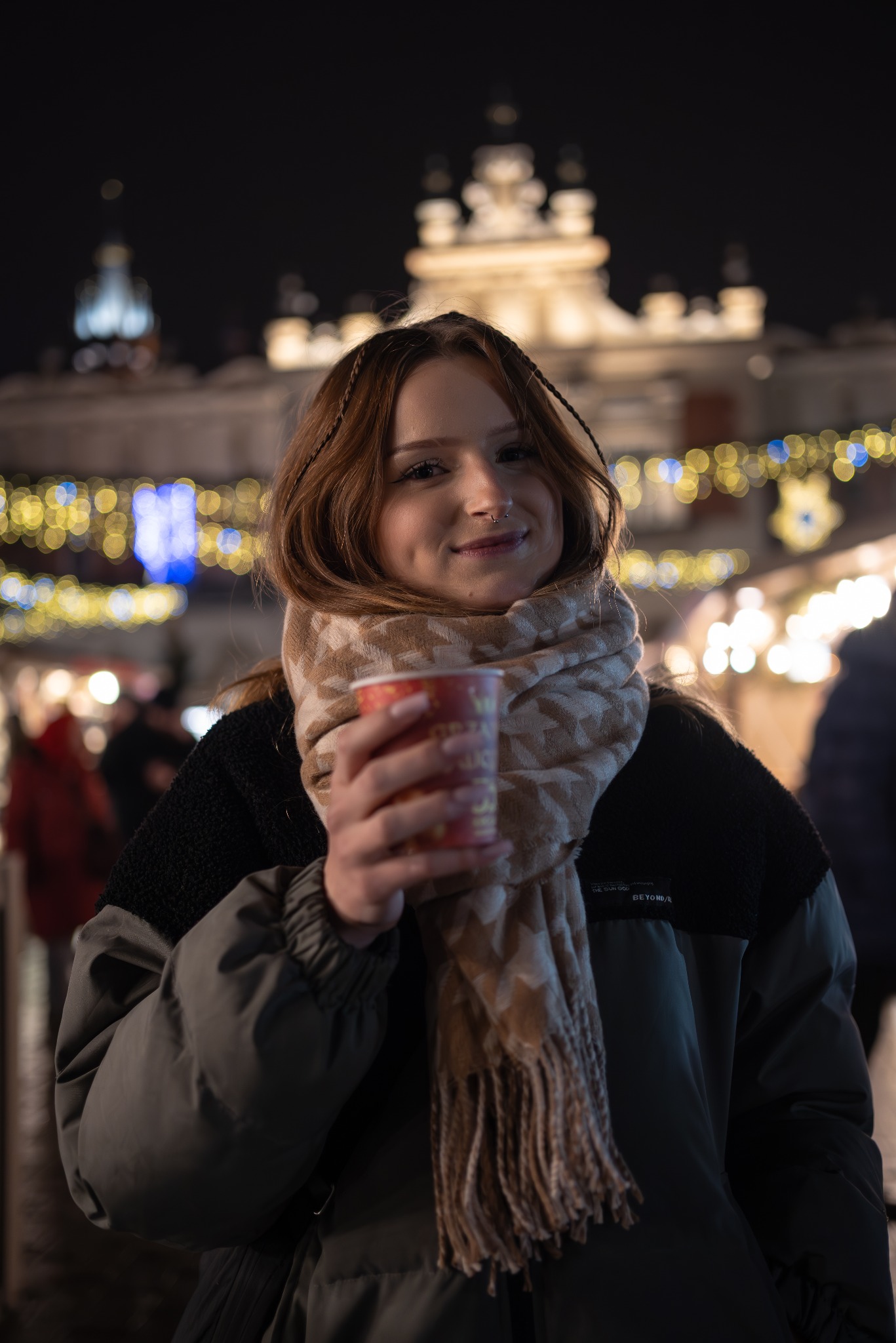 Uśmiechnięta dziewczyna z kolczykiem w nosie, w szaliku i kurtce na tle rozświetlonego świątecznego rynku, trzymająca kubek z napojem.