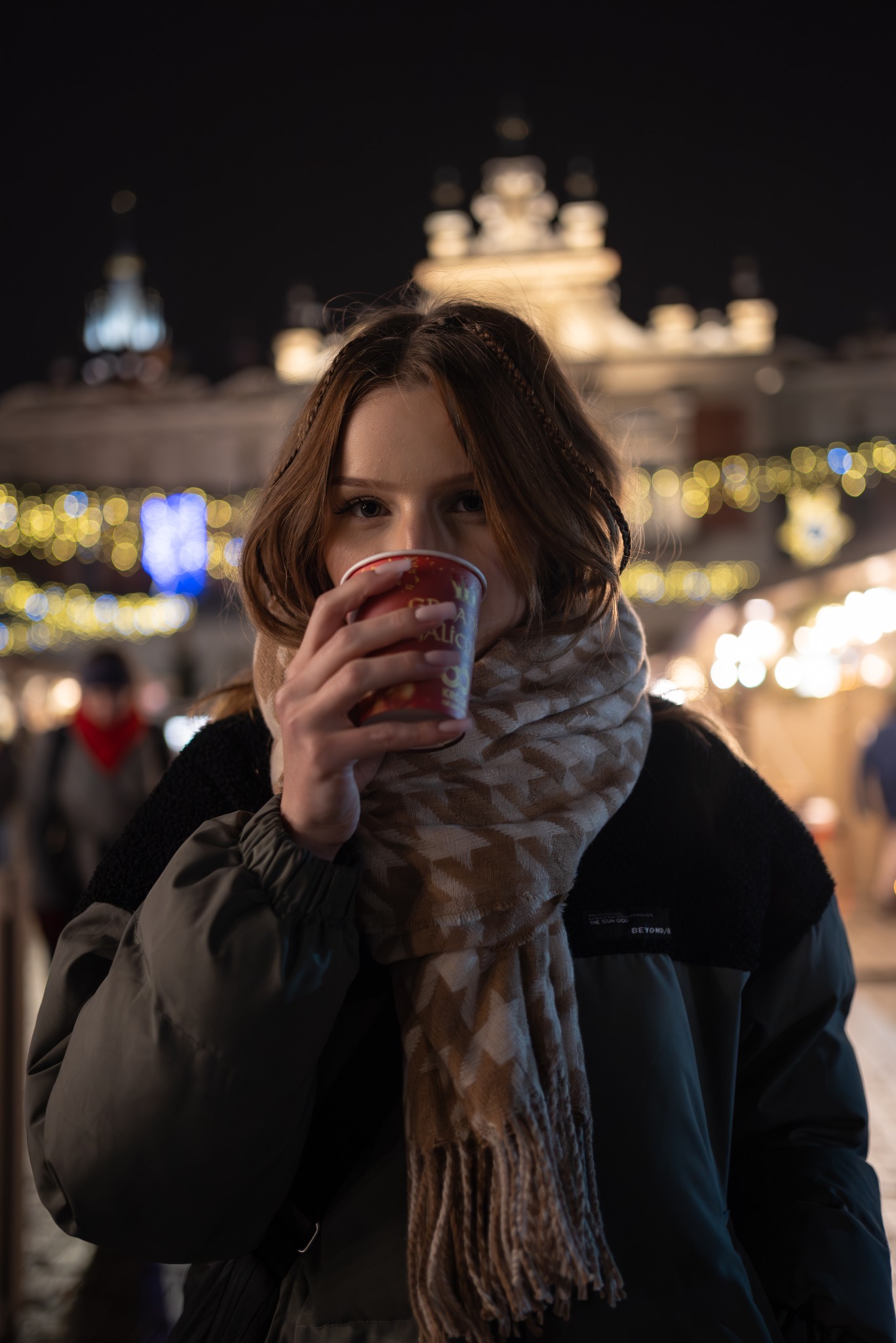 Młoda kobieta w Zabrzu pije grzane wino na tle rozświetlonego świątecznego jarmarku z zabytkową architekturą w tle, ubrana w zimową kurtkę i wzorzysty szal.
