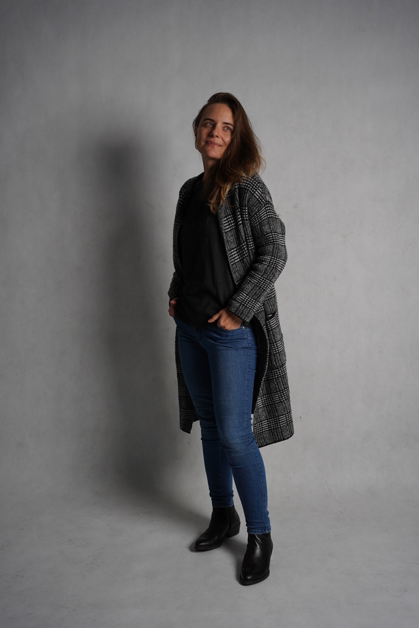 Kobieta w dżinsach, czarnej bluzce i płaszczu w kratę, pozująca na szarym tle, z rękami w kieszeniach i uśmiechem na twarzy.