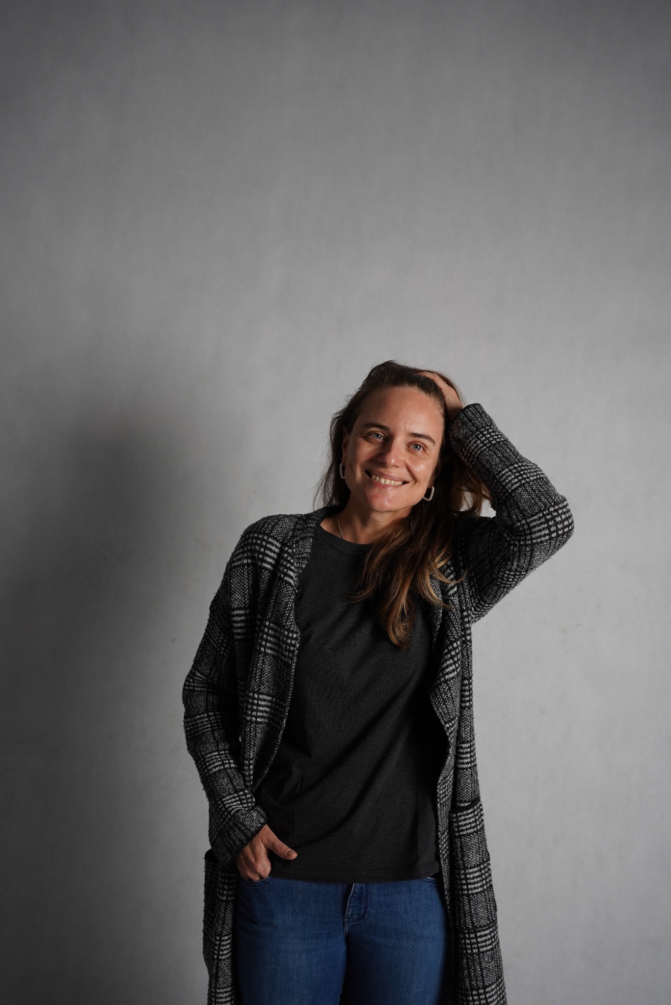 Uśmiechnięta kobieta w szarym swetrze i kraciastej narzutce pozuje na tle szarej ściany, dłoń we włosach, druga ręka w kieszeni dżinsów.
