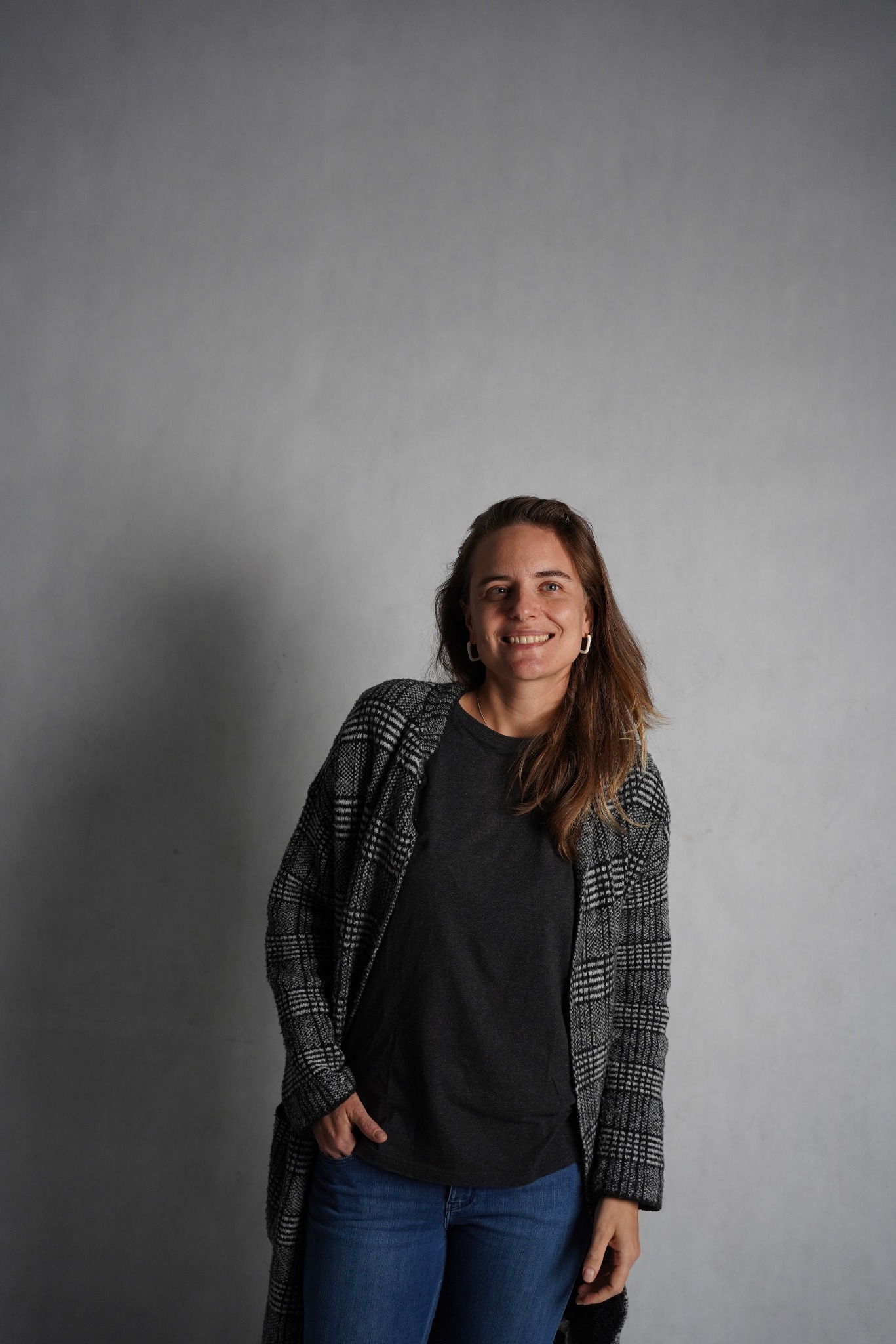 Portret uśmiechniętej kobiety w jeansach, grafitowej koszulce i kraciastej narzutce na szarym tle.