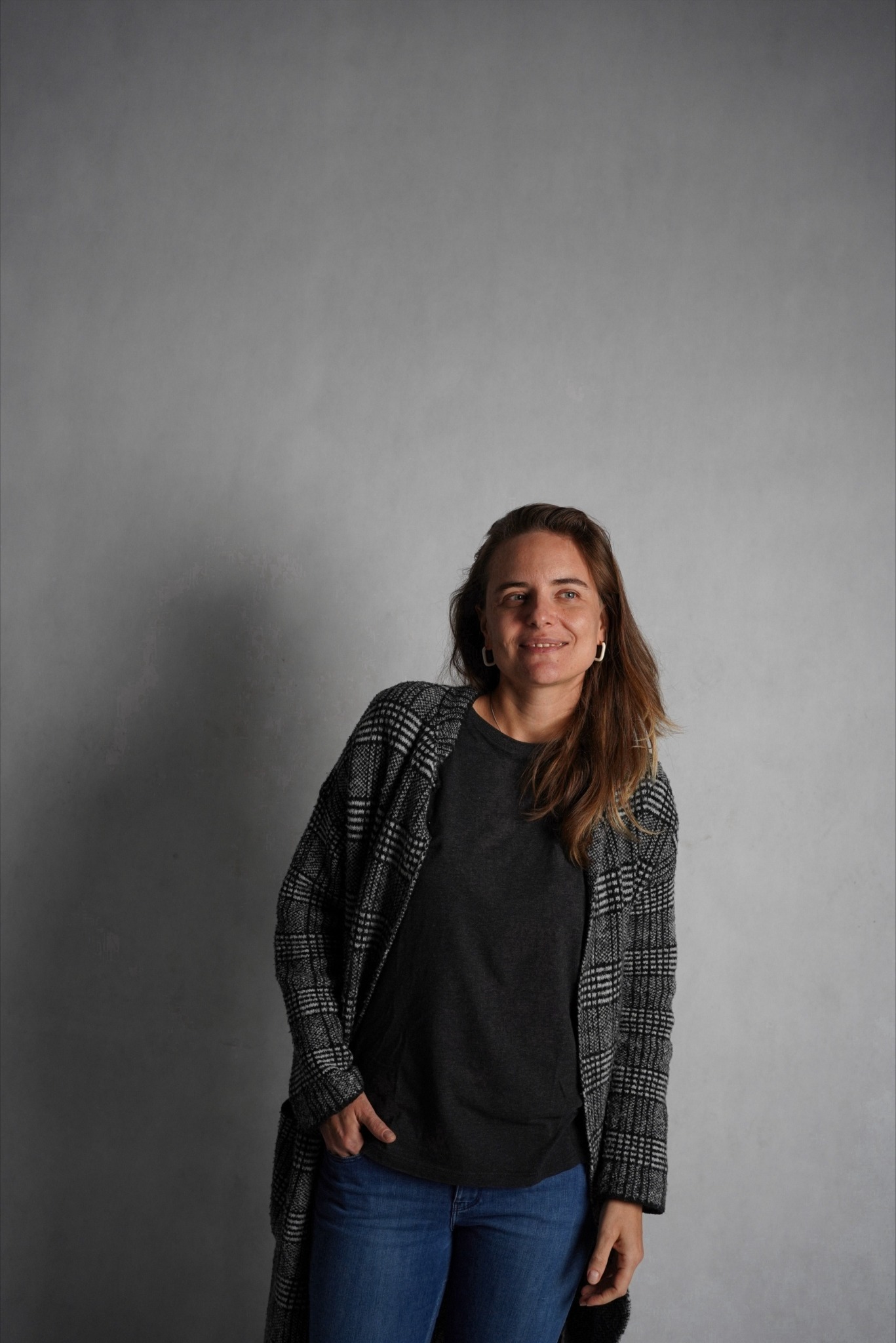 Portret uśmiechniętej kobiety w dżinsach i kraciastej narzutce na tle szarej ściany, z ręką w kieszeni.