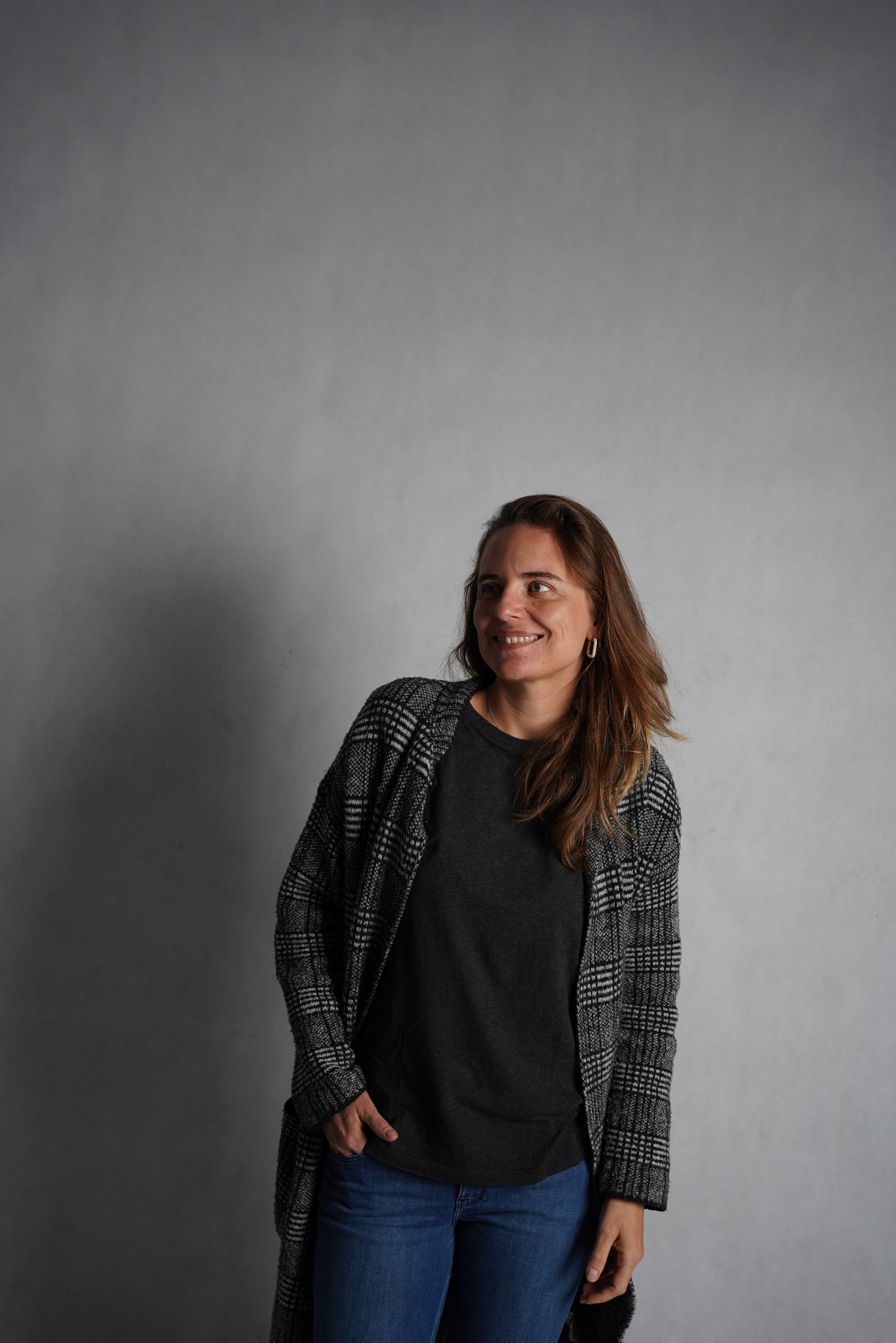 Kobieta z długimi brązowymi włosami, w dżinsach, czarnym T-shircie i szarym kardiganie w kratę, uśmiecha się i patrzy w bok na szarym tle.