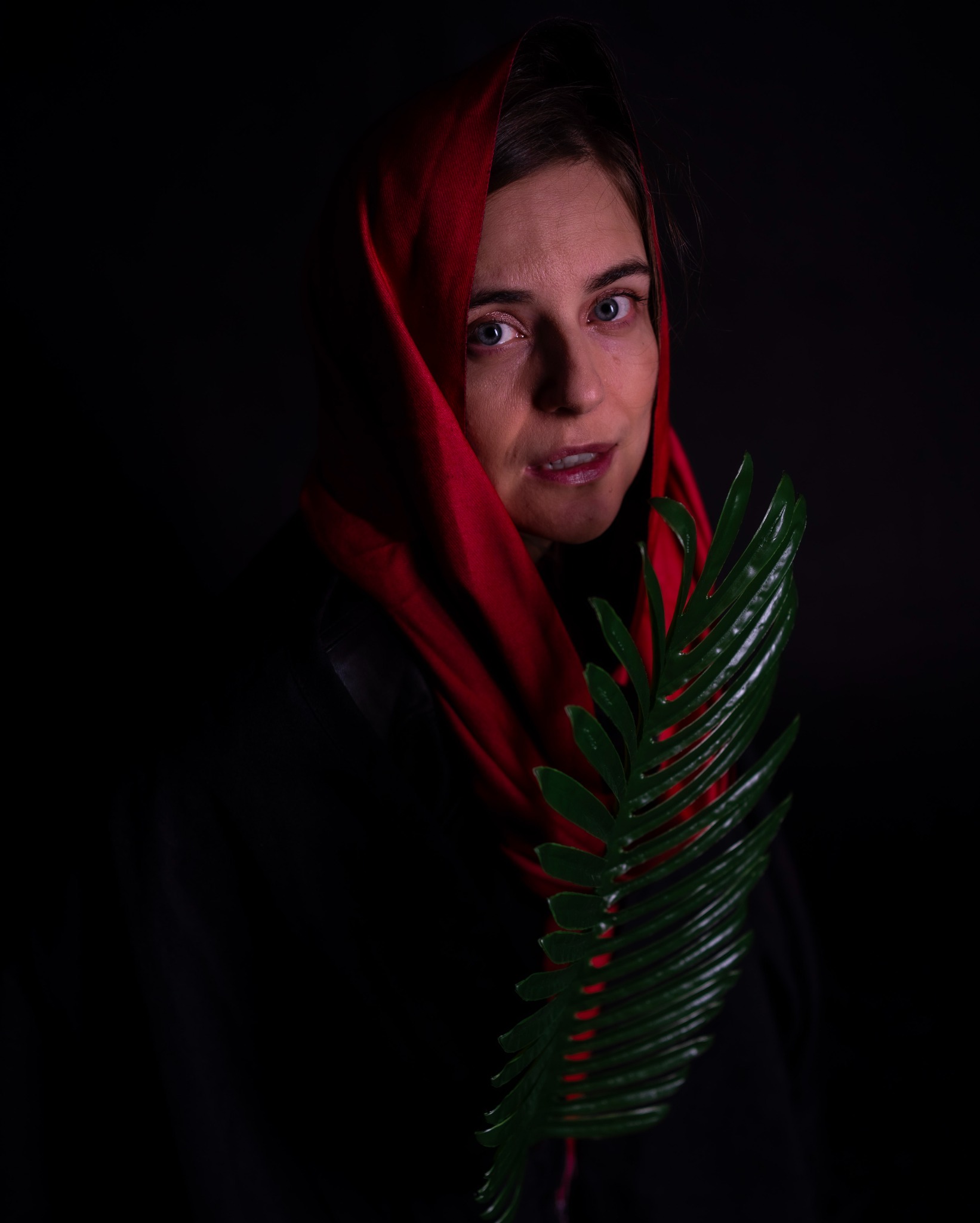 Portret kobiety z czerwonym szalem na głowie, trzymającej zieloną gałązkę na ciemnym tle.