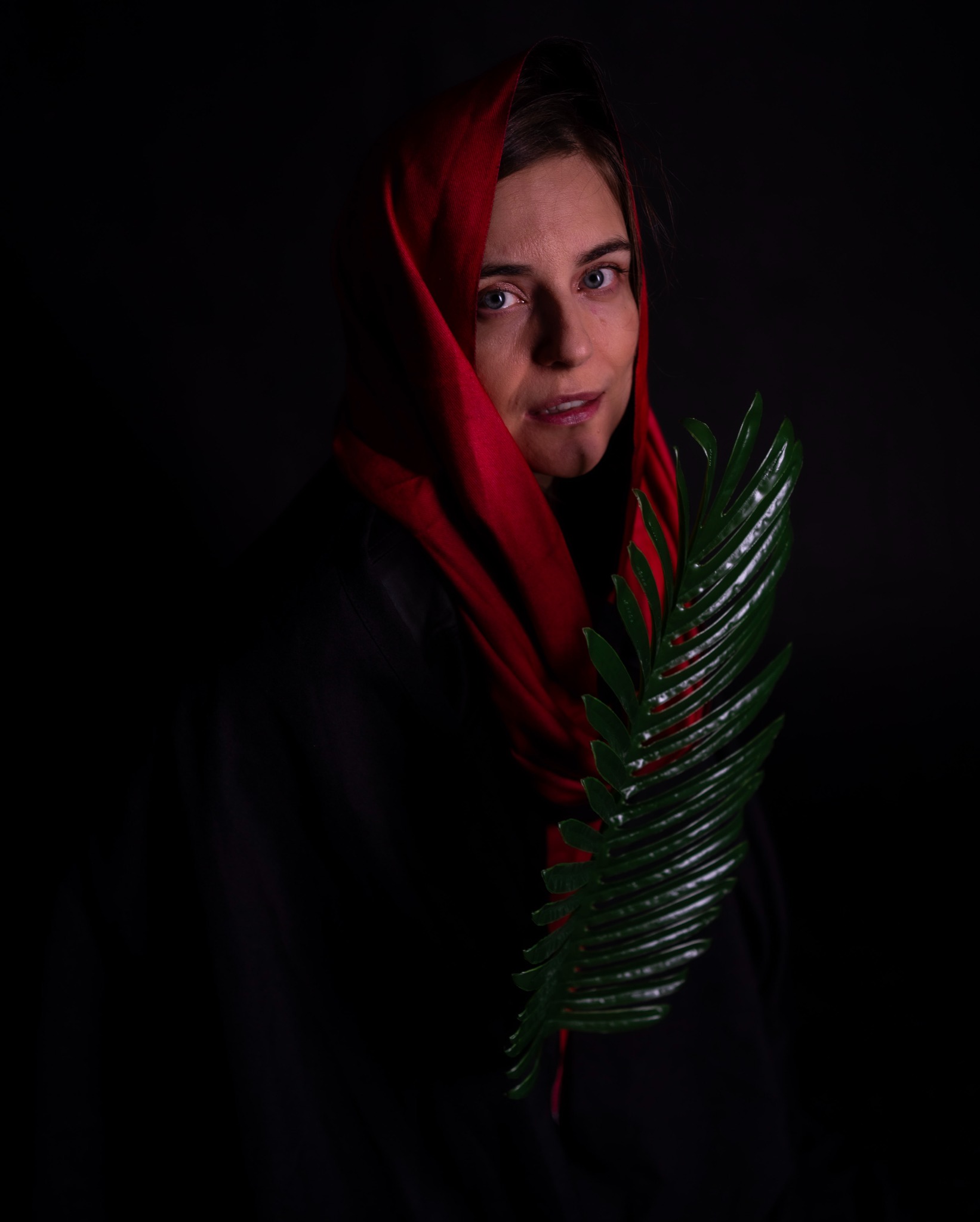 Portret kobiety z czerwonym szalem na głowie, trzymającej zielony liść na ciemnym tle.