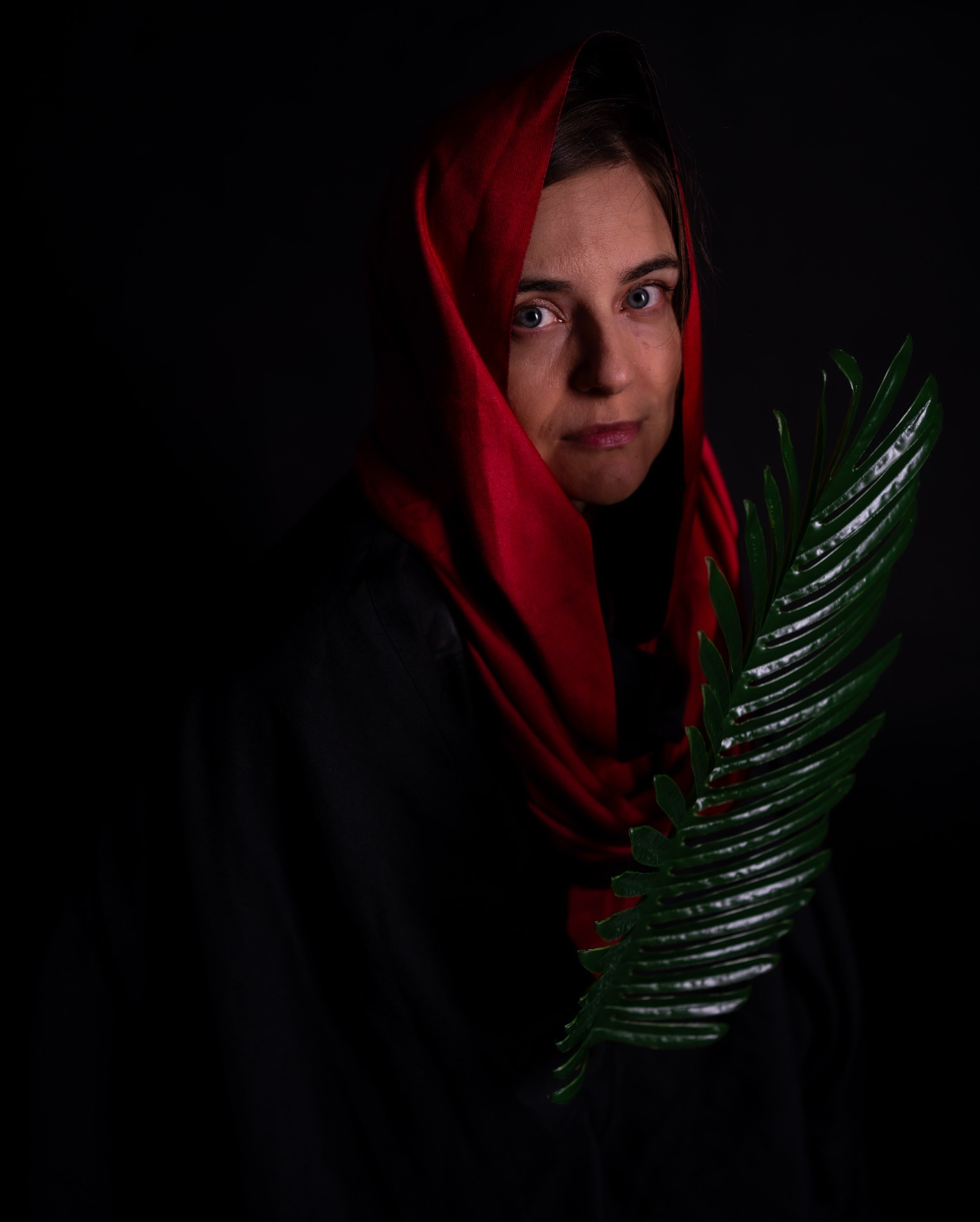 Portret kobiety z czerwonym nakryciem głowy i zielonym liściem na ciemnym tle.