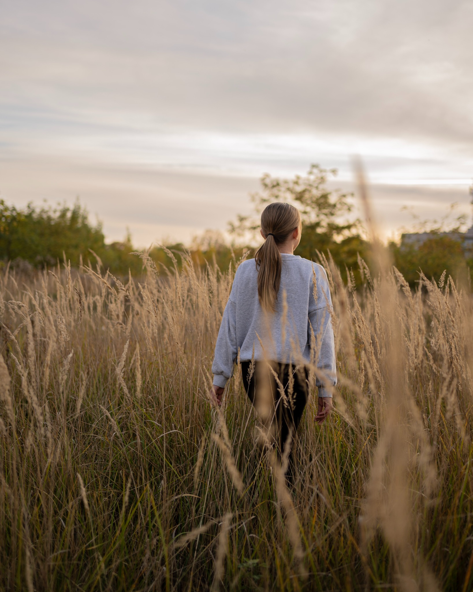 Kobieta z włosami związanymi w kucyk, ubrana w szarą bluzę, idzie przez pole wyschniętej trawy w kierunku zachodzącego słońca.