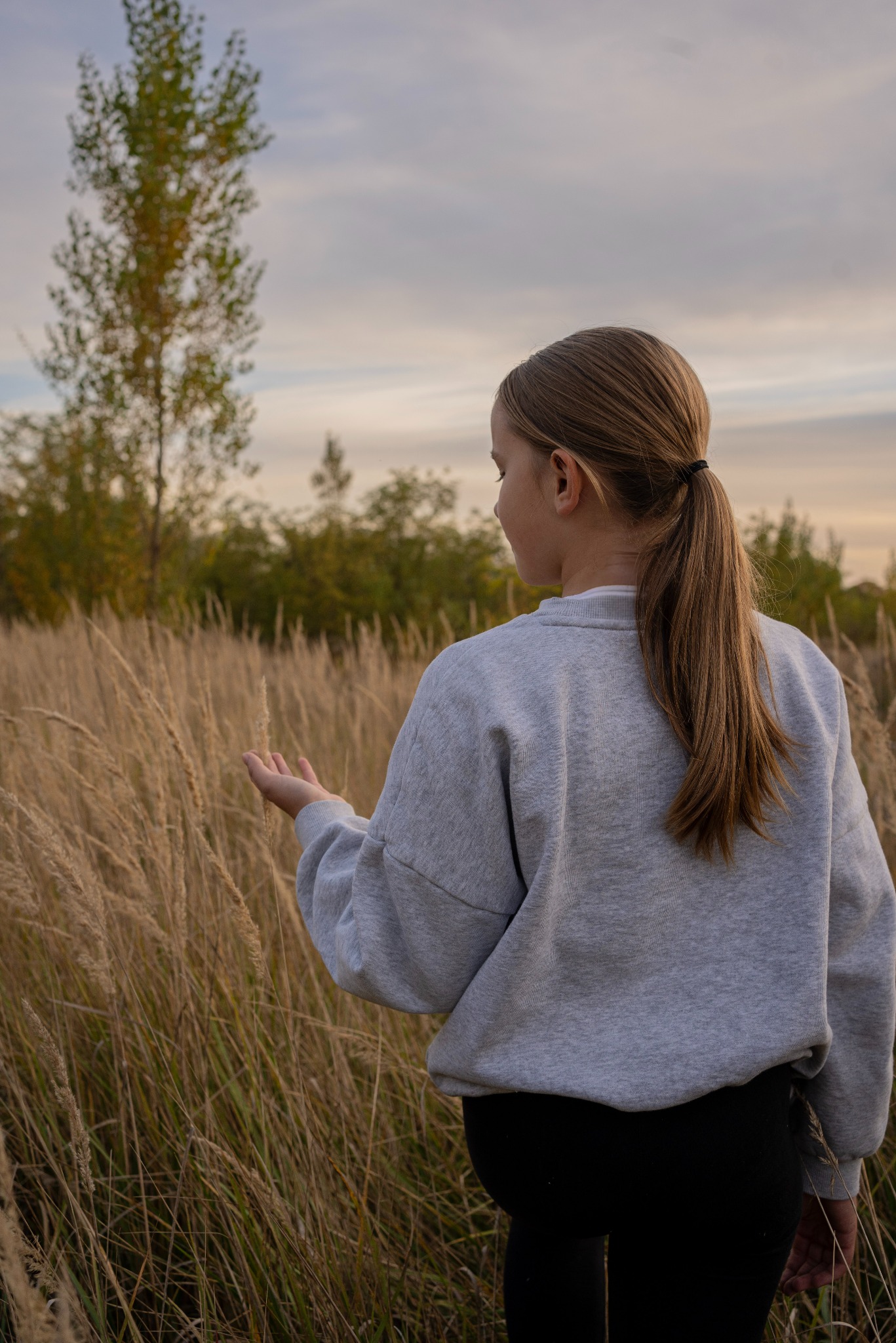 Dziewczynka z długim kucykiem w szarej bluzie dotyka trawy na łące o zachodzie słońca.