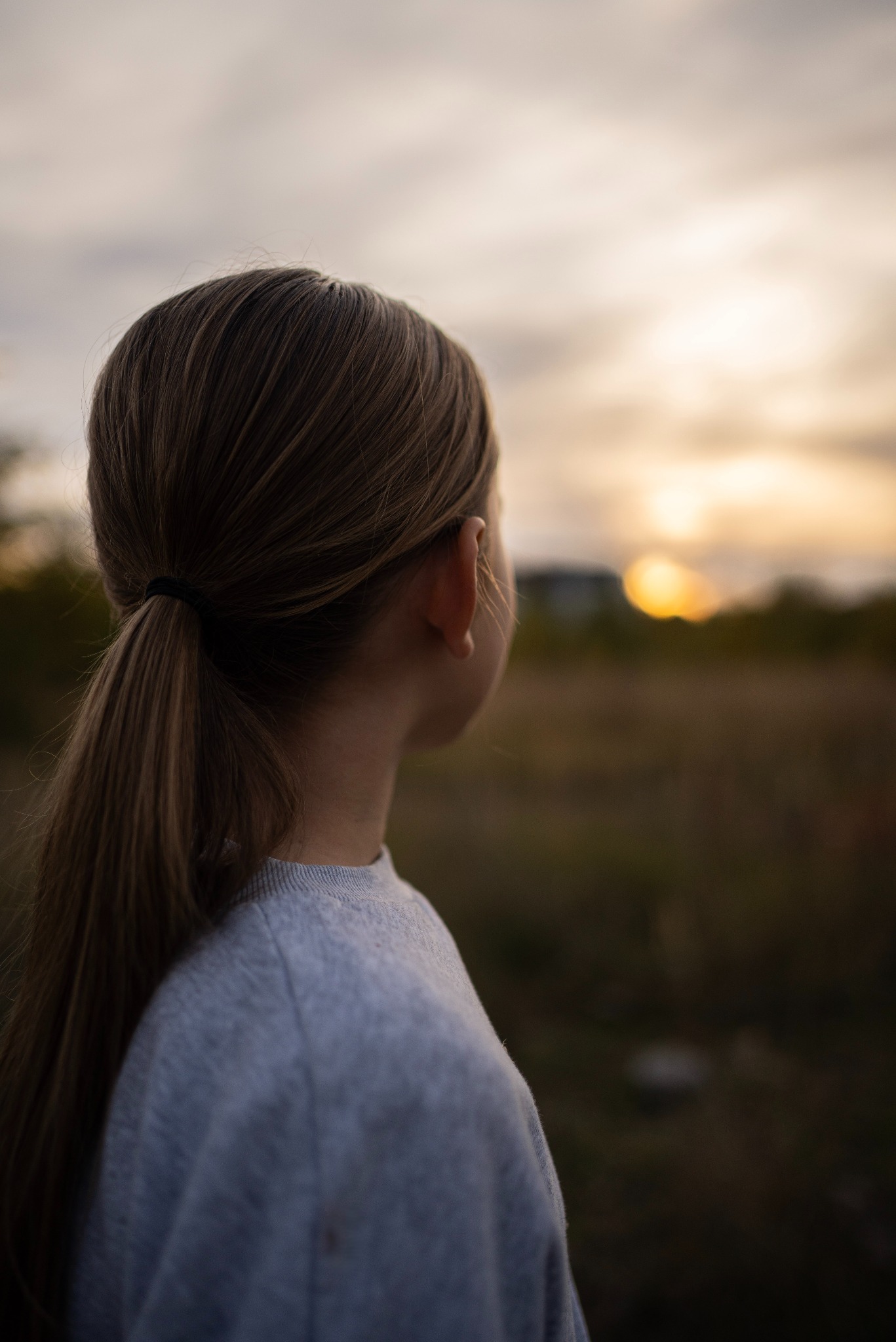 Dziewczynka z blond włosami spiętymi w kucyk patrzy w dal na zachodzące słońce nad polami.