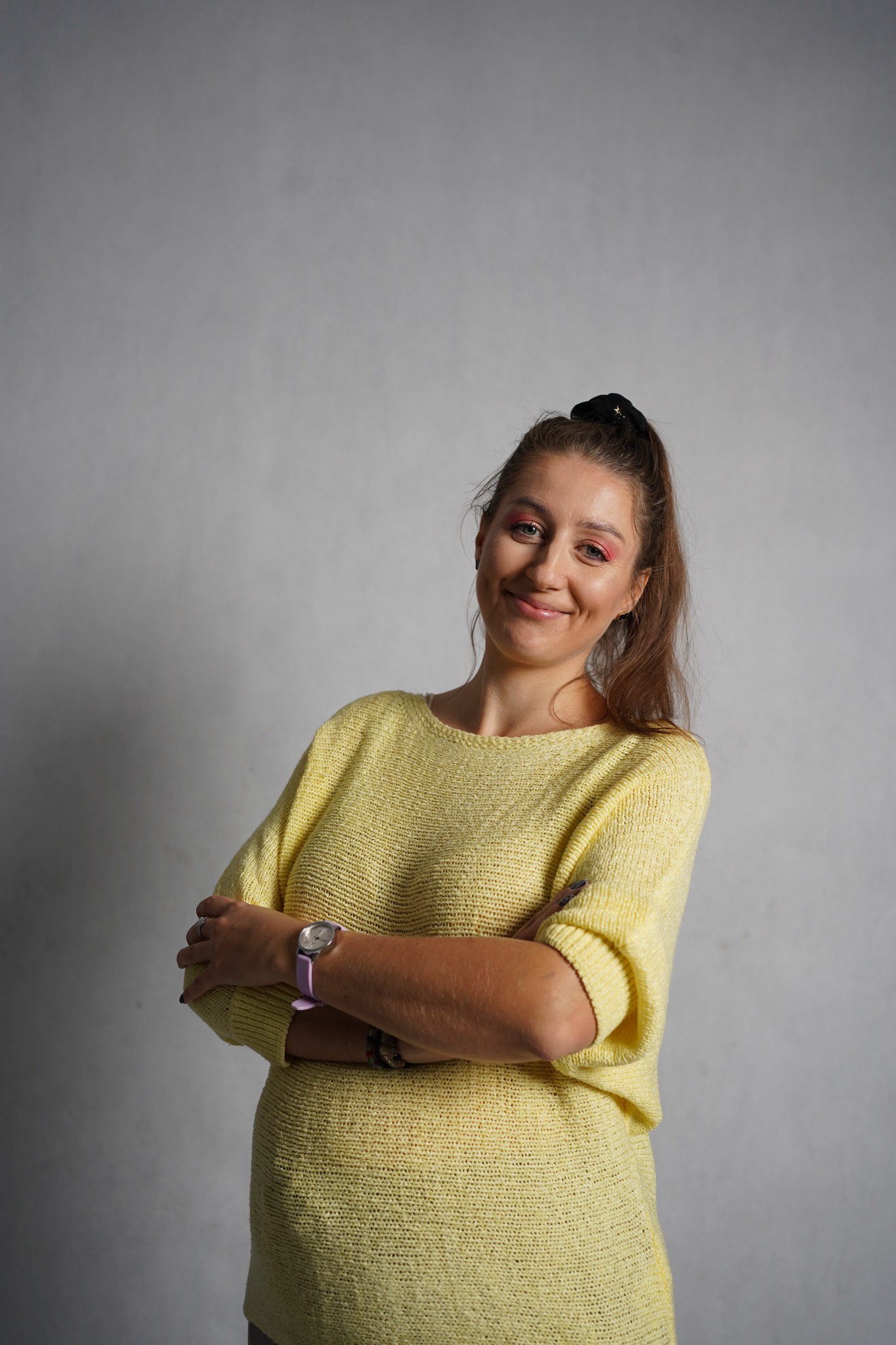 Portret uśmiechniętej kobiety w żółtym swetrze, z rękami skrzyżowanymi na piersiach, z zegarkiem na ręce i włosami upiętymi w kucyk, na jasnoszarym tle.