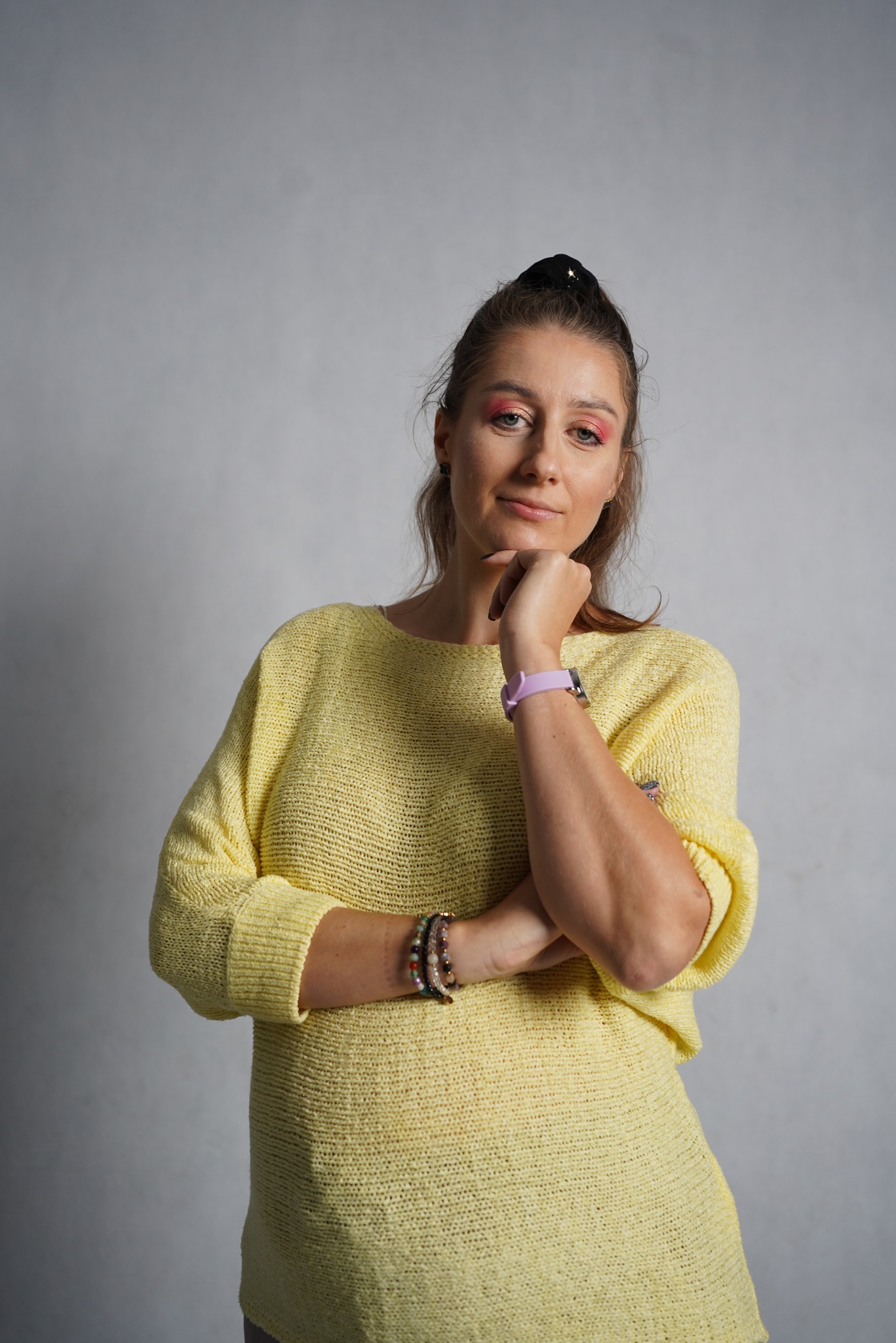 Portret kobiety w żółtym swetrze z rękami skrzyżowanymi pod brodą, pastelowy makijaż oczu, bransoletki na nadgarstku, szare tło.