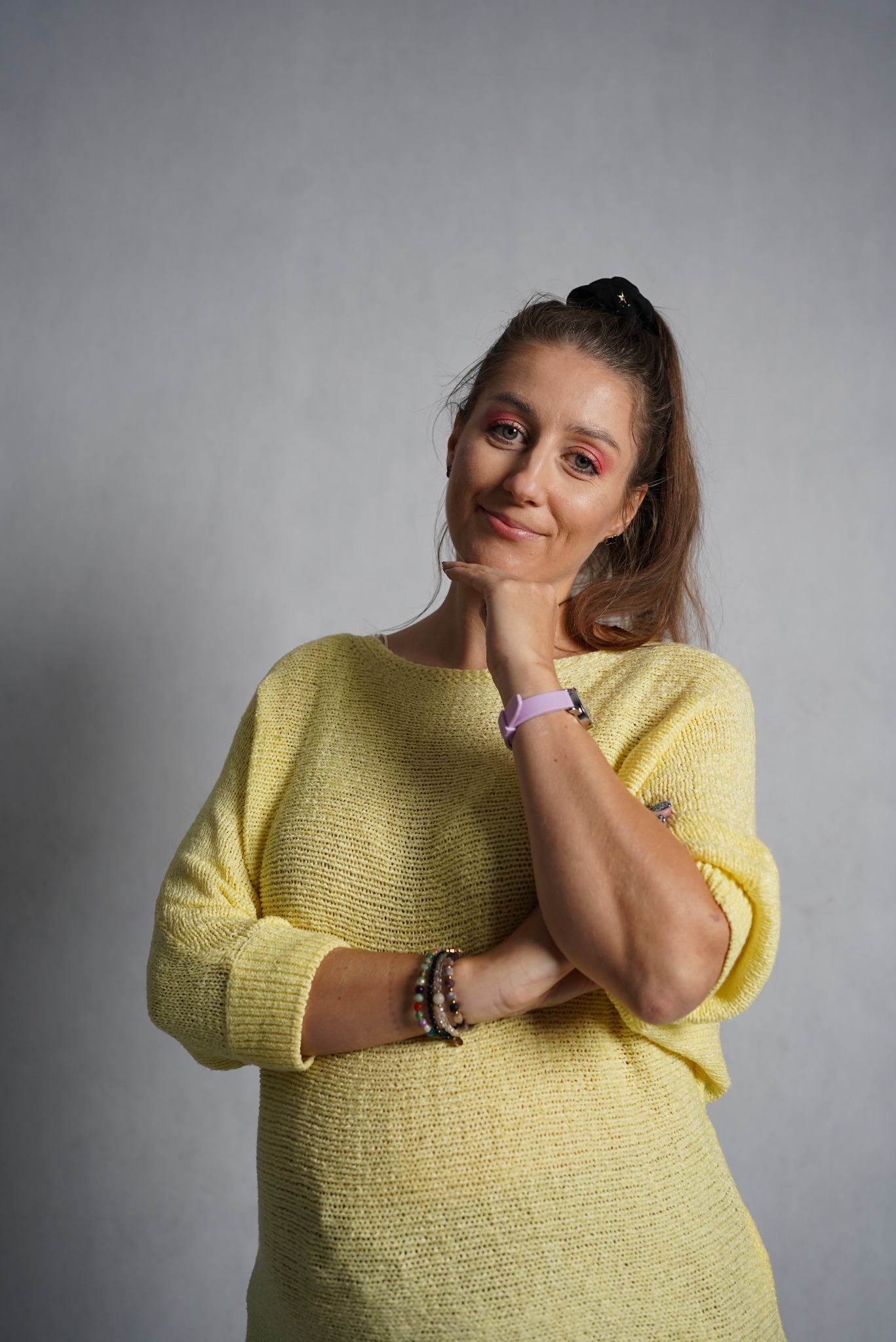 Portret uśmiechniętej kobiety w żółtym swetrze, z ręką pod brodą, z widocznymi bransoletkami z kamieni i fioletowym smartwatchem.