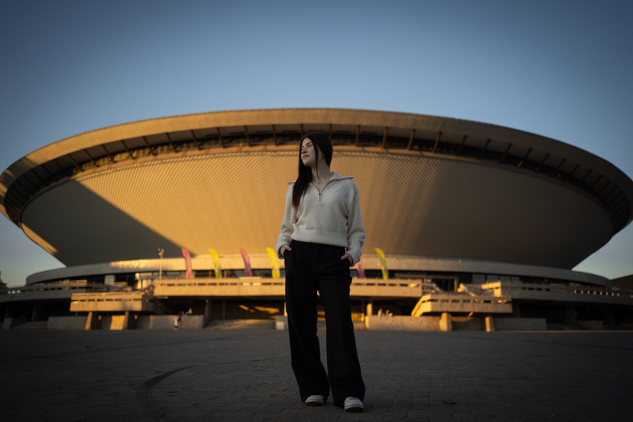 Młoda kobieta w swetrze i szerokich spodniach pozuje na tle Hali Widowiskowo-Sportowej Spodek w Katowicach podczas zachodu słońca.