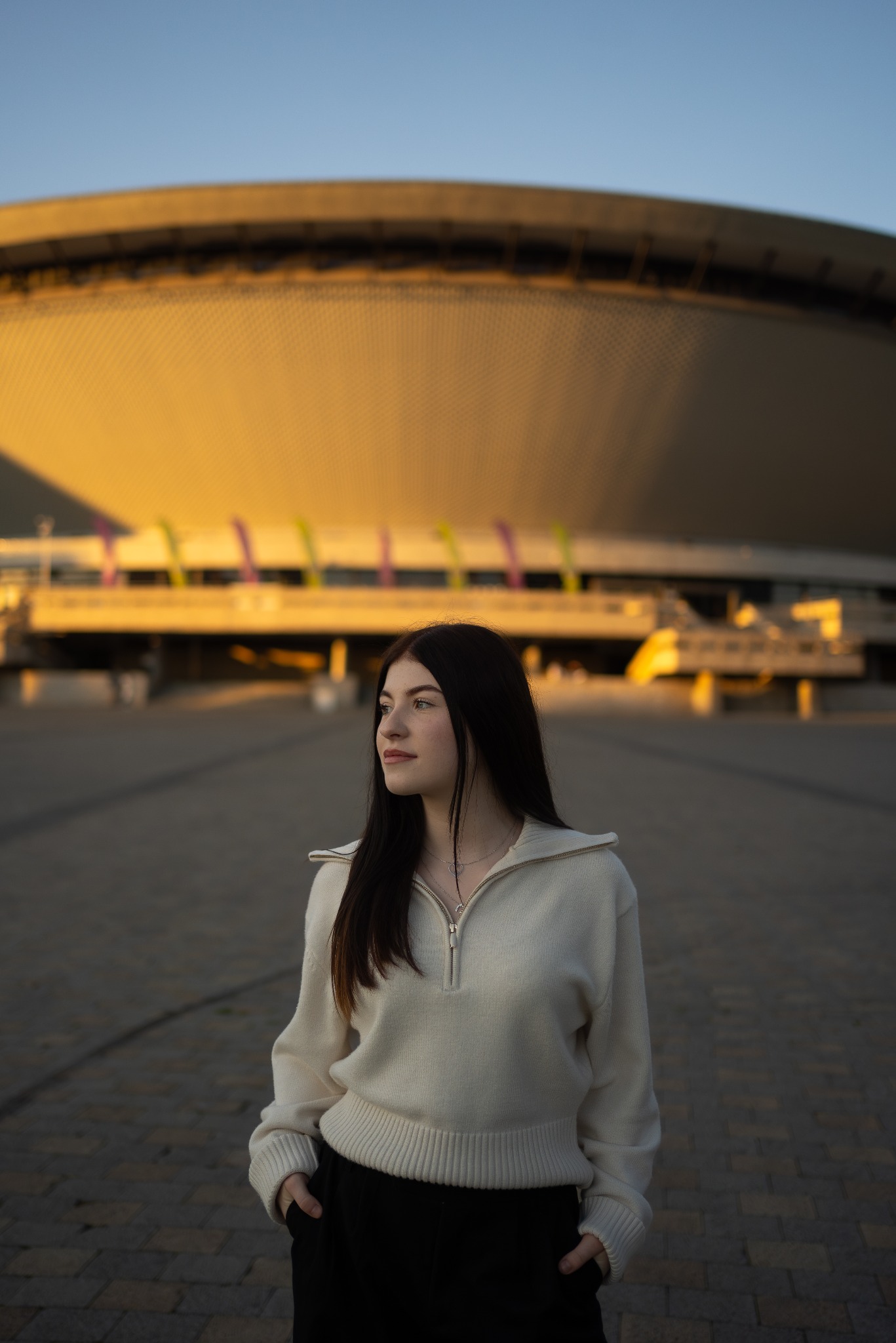Portret młodej kobiety w kremowym swetrze na tle Stadionu Śląskiego oświetlonego zachodzącym słońcem.