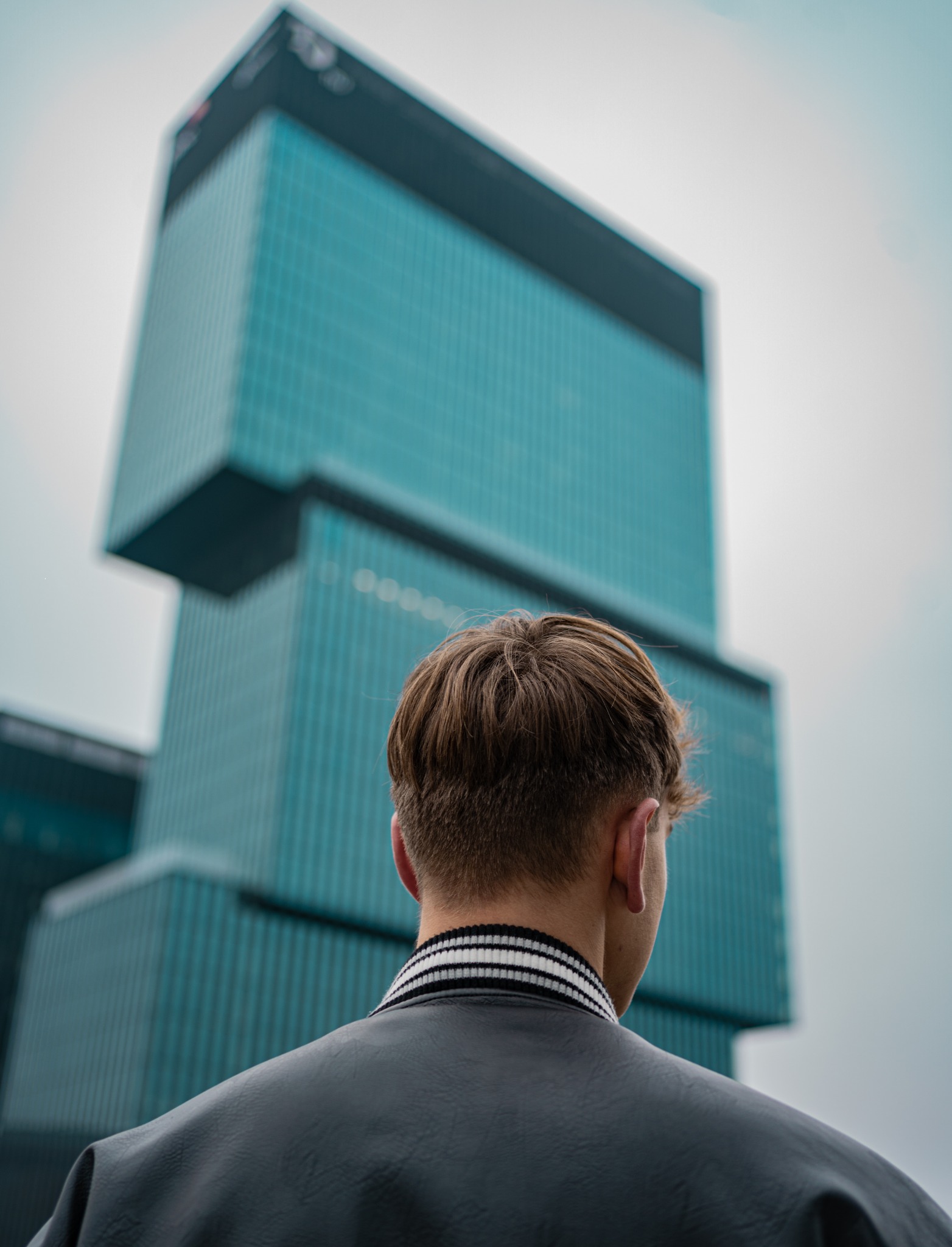 Mężczyzna w kurtce bomber patrzy na nowoczesny biurowiec o szklanej fasadzie w odcieniach błękitu, z widocznym karkiem i tyłem głowy.