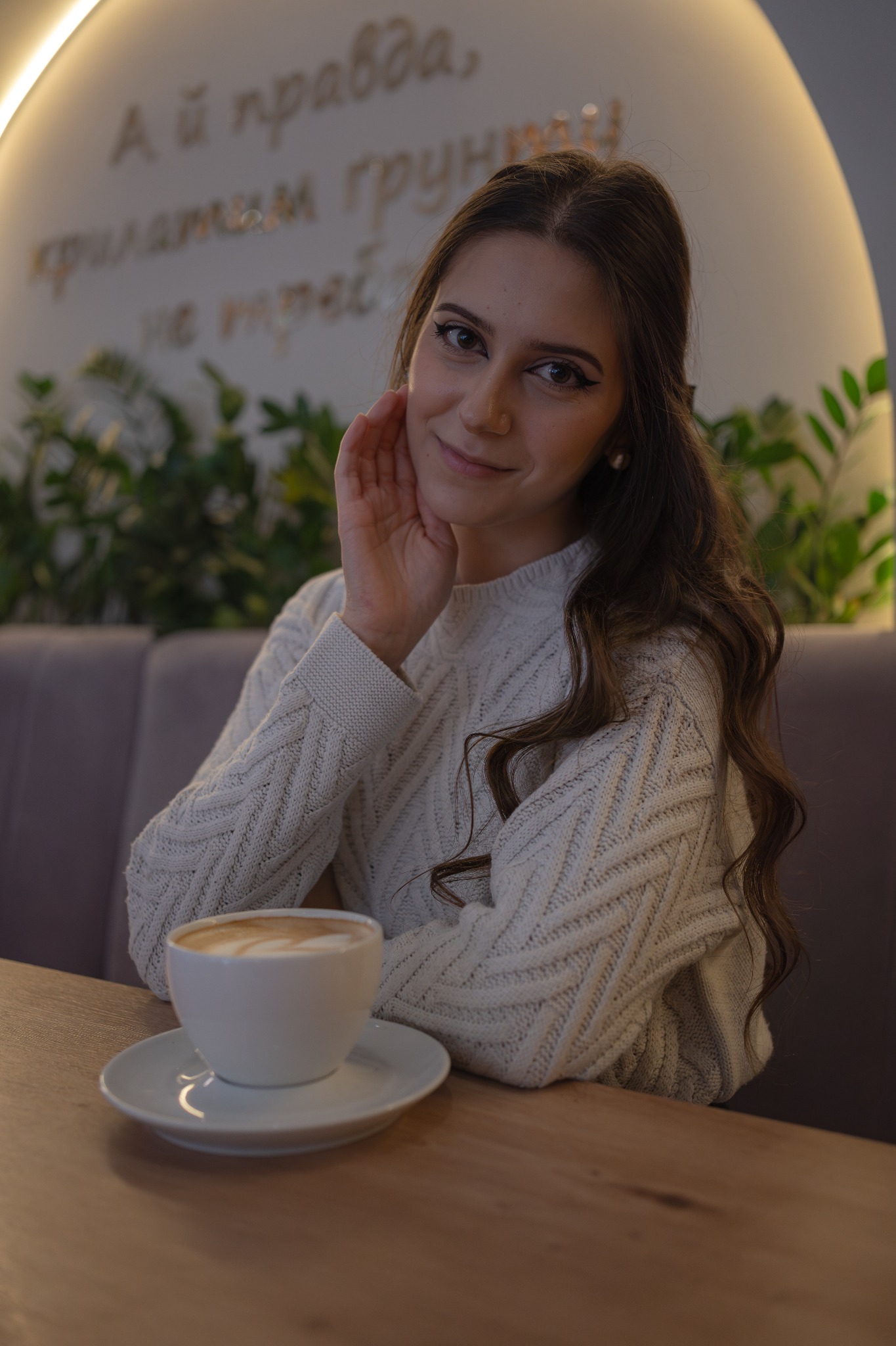 Uśmiechnięta brunetka w swetrze z kawą latte w kawiarni z napisem na ścianie w tle.