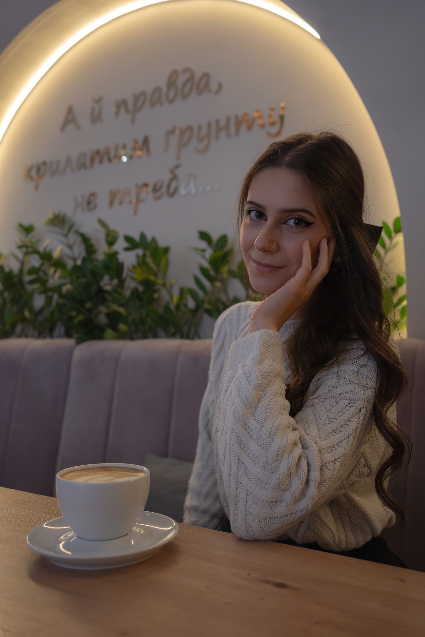 Uśmiechnięta kobieta w swetrze siedzi przy stoliku w kawiarni z filiżanką cappuccino, w tle dekoracyjne oświetlenie i rośliny.