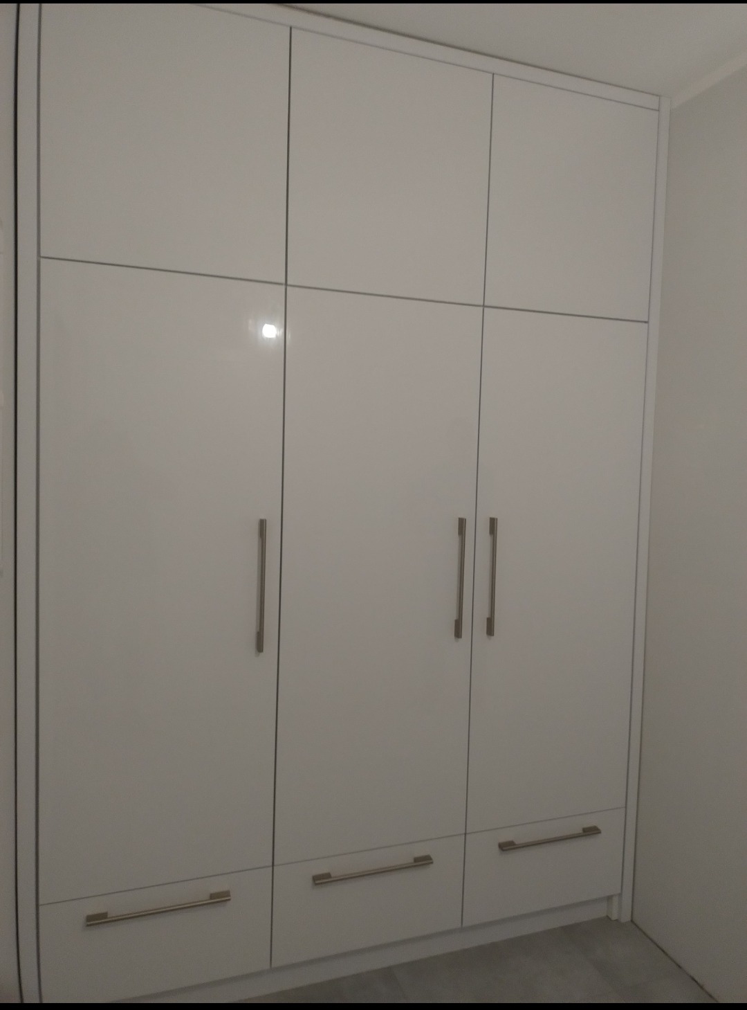 Biała, lakierowana szafa wnękowa z trzema parami drzwi i trzema szufladami na dole, widoczne metalowe uchwyty.