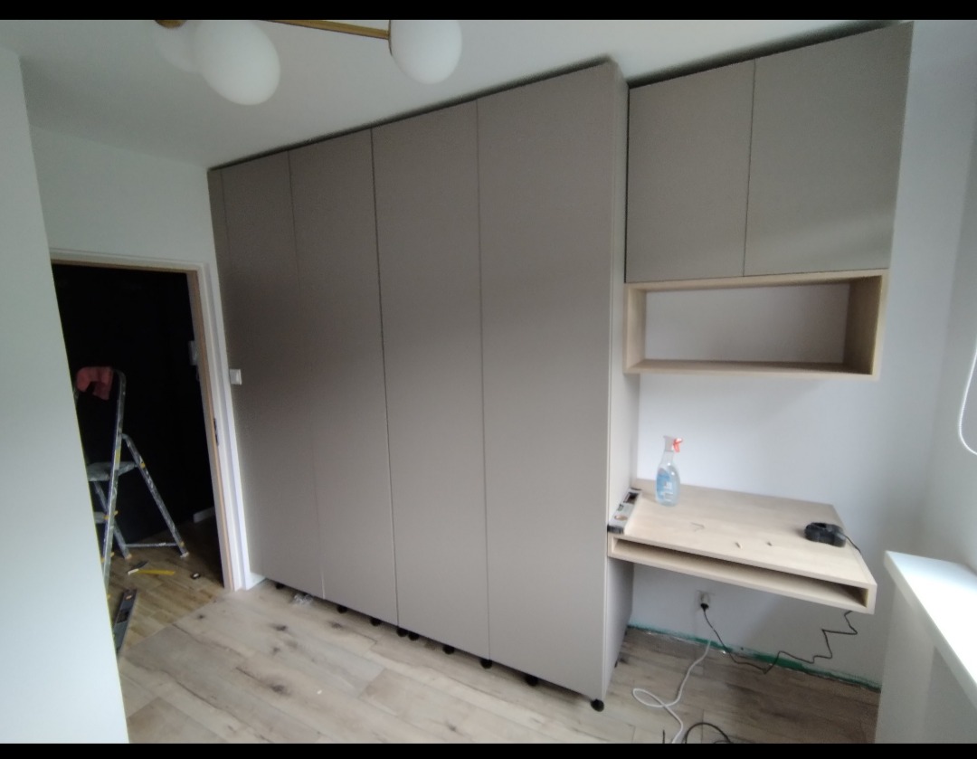 Szara zabudowa meblowa w pokoju z biurkiem i szafkami wiszącymi, widoczna drabina w tle, podłoga z paneli.