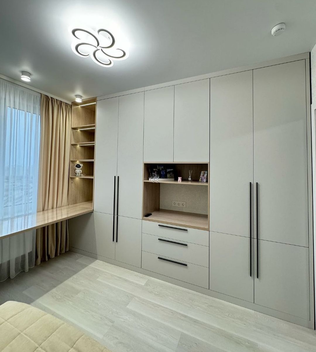 Wnętrze pokoju z zabudową meblową: szafa z szufladami, otwartymi półkami i biurkiem przy oknie, wykończona w jasnych odcieniach szarości i drewna, oświetlona nowoczesną lampą sufitową.