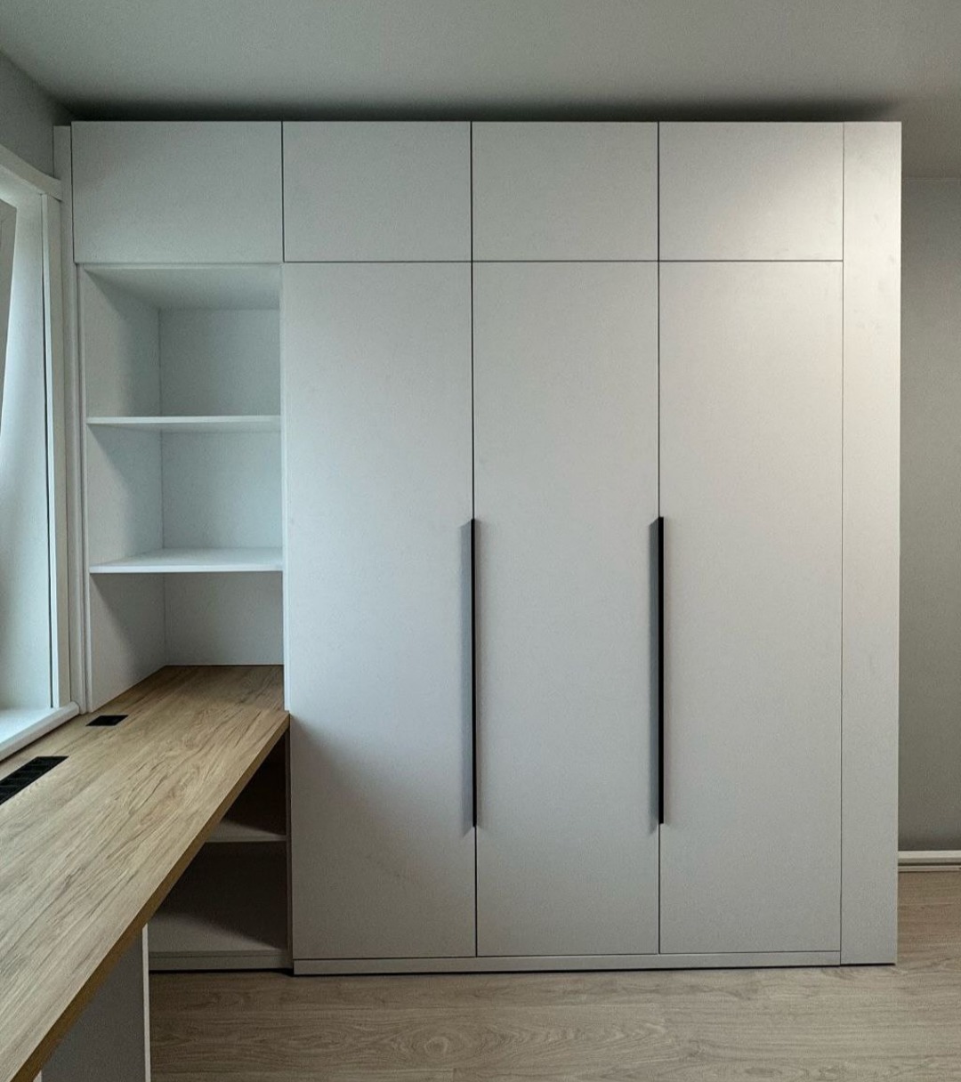 Biała zabudowa meblowa z szafą i biurkiem z drewnianym blatem, minimalistyczny design, czarne uchwyty, jasne wnętrze.