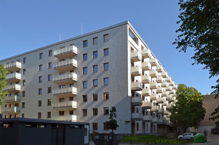 Nowoczesny, biały budynek mieszkalny z licznymi balkonami z przezroczystymi balustradami, widziany z poziomu ulicy w słoneczny dzień. Na pierwszym planie widoczne kontenery na śmieci i fragment...