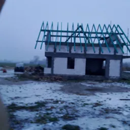 Surowy stan dwukondygnacyjnego domu w trakcie budowy z niebieskimi krokwiami dachowymi, widoczny pracownik w odblaskowej kamizelce w tle, zimowa sceneria z resztkami śniegu.