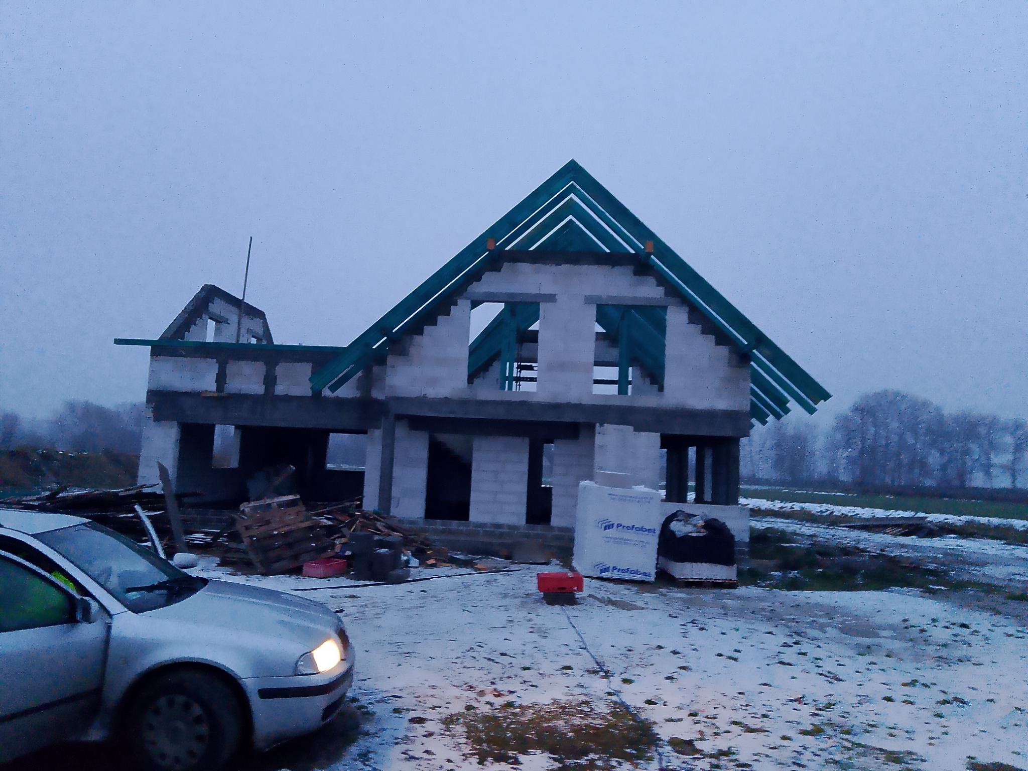 Surowy stan domu w trakcie budowy z elementami więźby dachowej w kolorze zielonym, widoczne bloczki betonowe i materiały budowlane na zewnątrz, samochód osobowy z włączonym reflektorem na pierwszym...