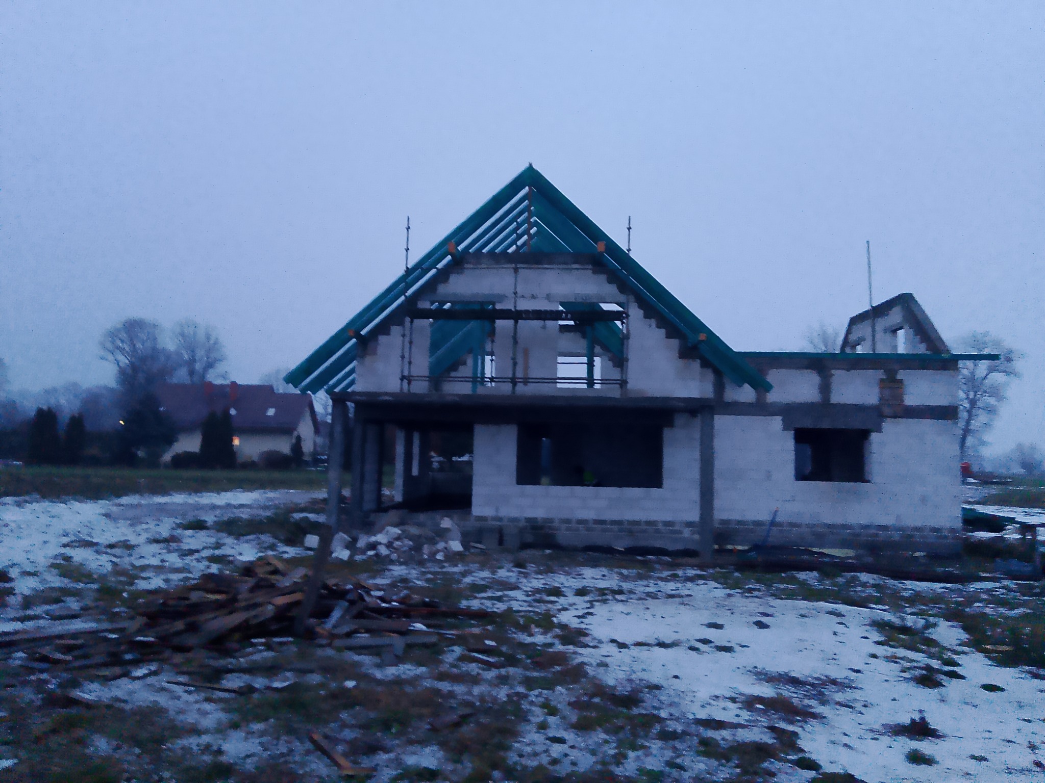Surowy stan domu murowanego w trakcie budowy, widoczne ściany z pustaków, konstrukcja dachu w kolorze zielonym, teren wokół pokryty resztkami śniegu, pochmurna pogoda.