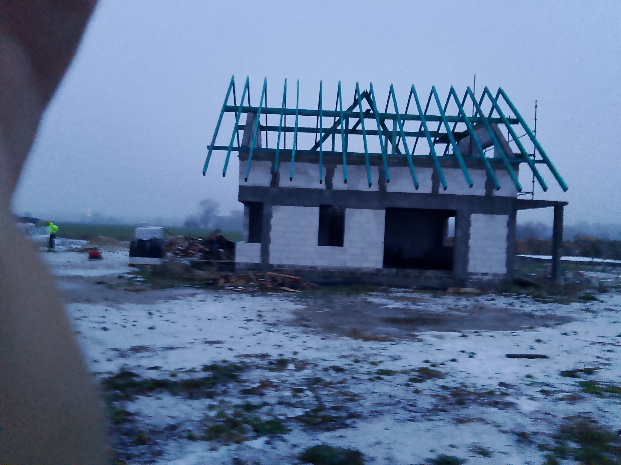 Surowy stan dwukondygnacyjnego domu w trakcie budowy z niebieskimi krokwiami dachowymi, widoczny pracownik w odblaskowej kamizelce w tle, zimowa sceneria z resztkami śniegu.