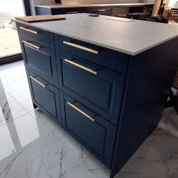 Renewood Piotr Mosór - Granatowa wyspa kuchenna z białym blatem marmurowym i złotymi uchwytami, na tle marmurowej podłogi i składanych drewnianych krzeseł.